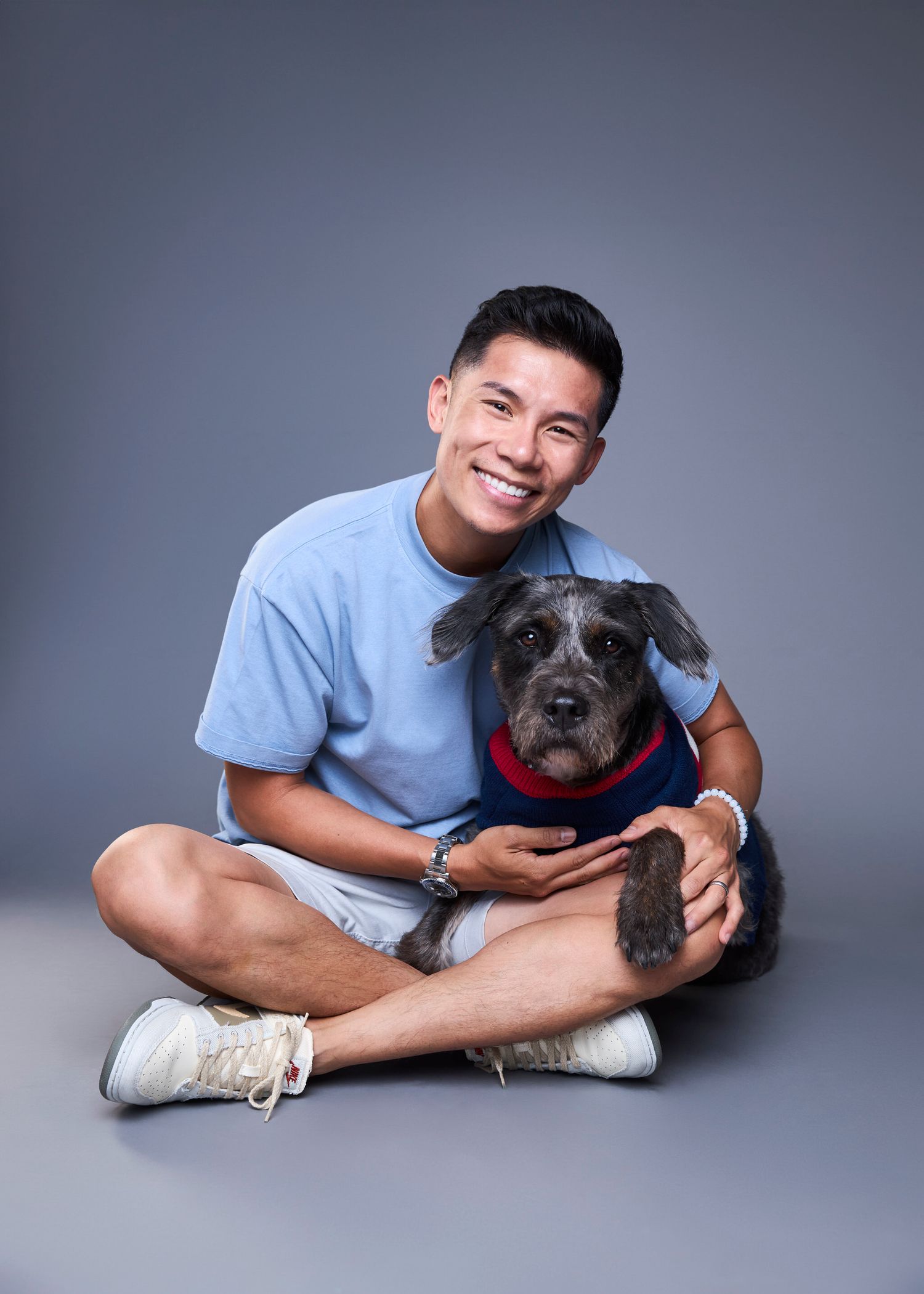 Young man in blue t-shirt sitting cross-legged on floor hugging a gray schnauzer dog wearing a red sweater.