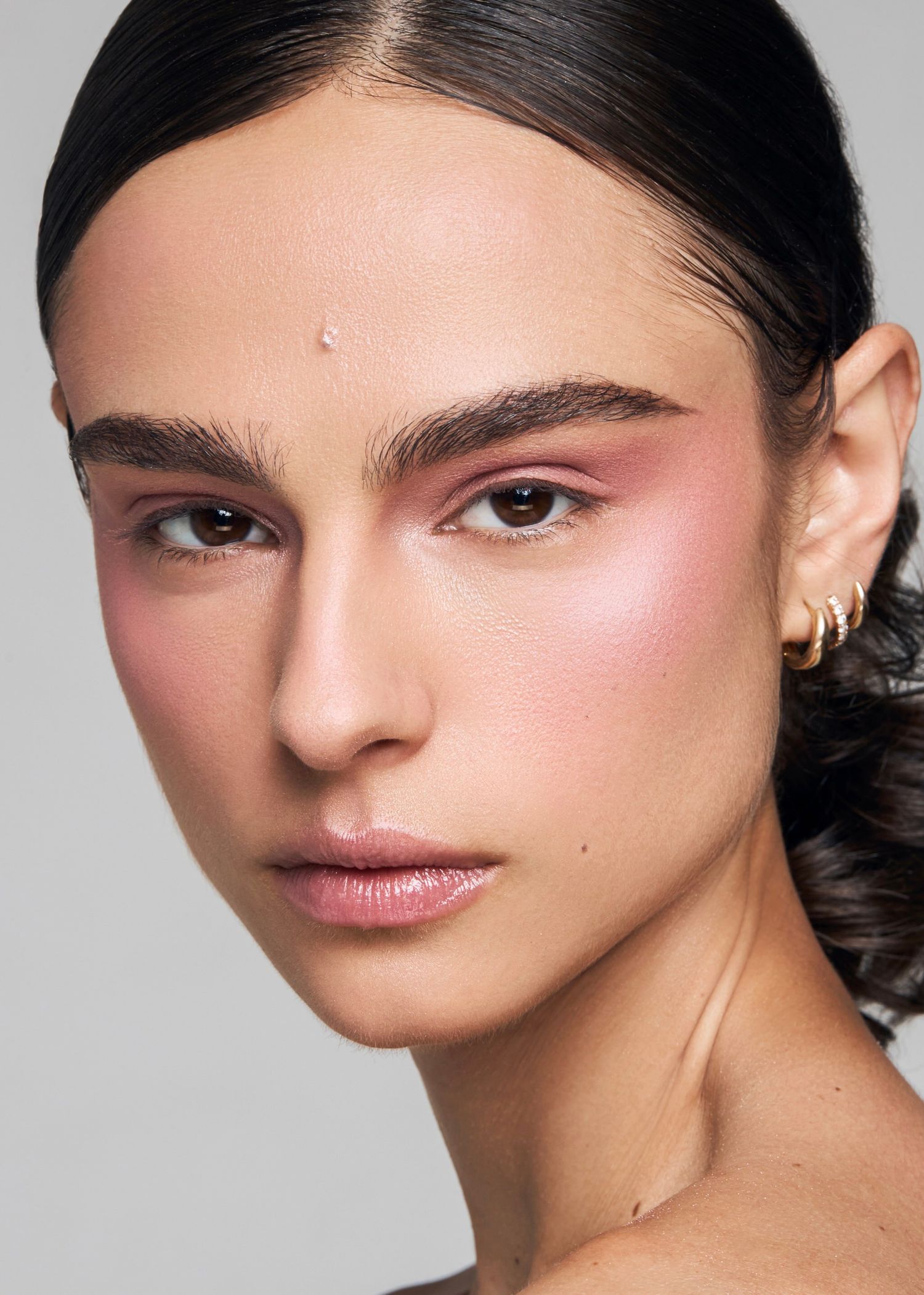 Close-up of a model wearing pink eye makeup, rosy blush, and gold hoop earrings against a grey background.