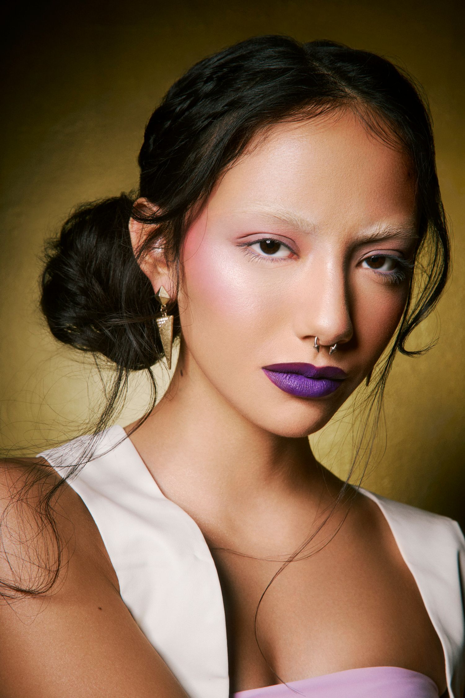 Close-up portrait of woman with purple lipstick, pink eyeshadow, nose ring, and upswept dark hair.