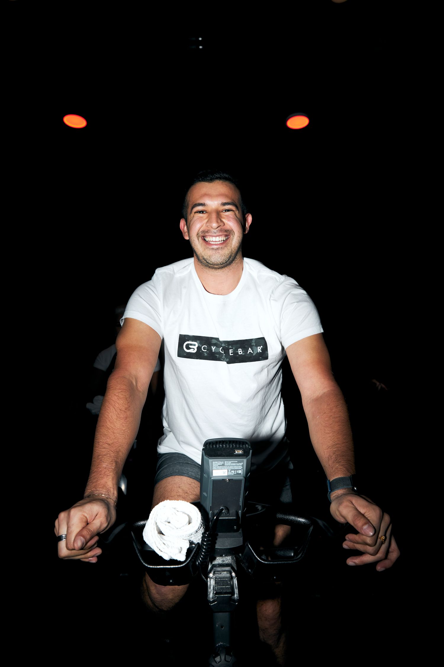 Smiling man riding a stationary bike in a white CycleBar t-shirt in a dark studio.