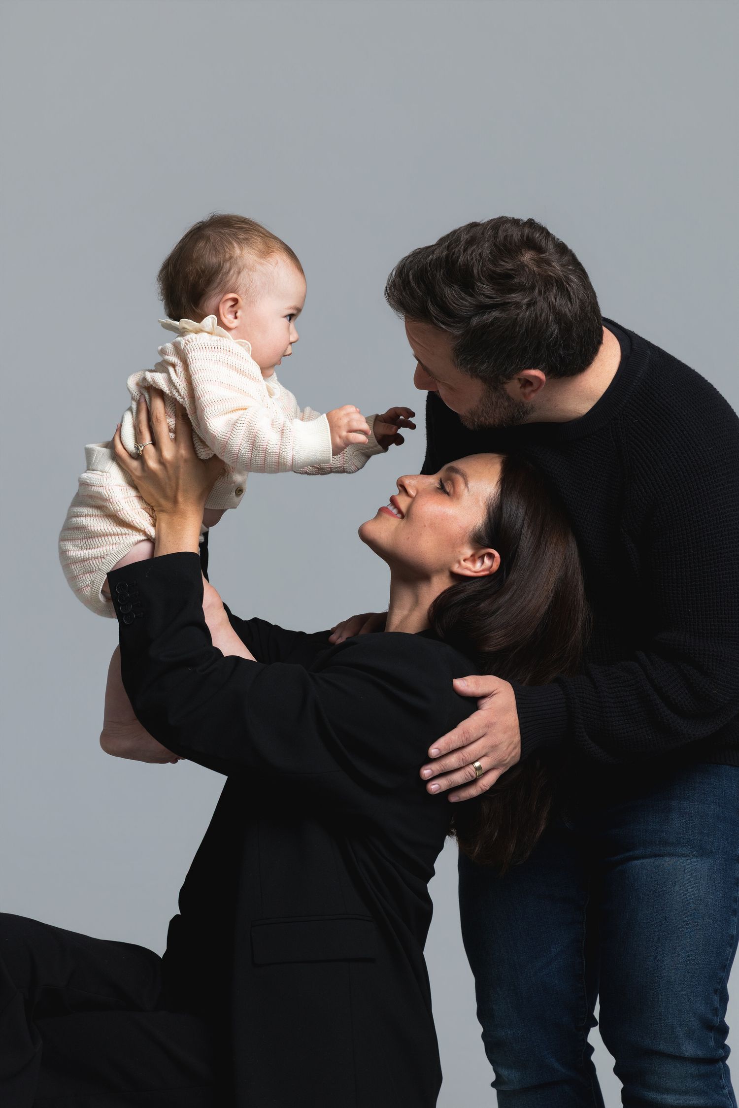 Happy family portrait with parents in black holding smiling baby in white outfit against gray background.