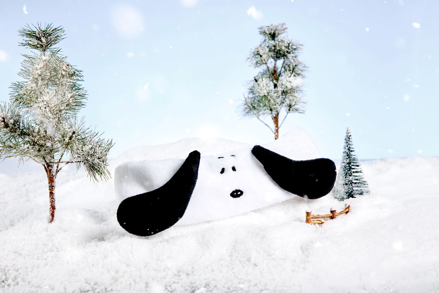 Cartoon dog toy lying in snow surrounded by snow-covered pine trees in a winter wonderland scene.