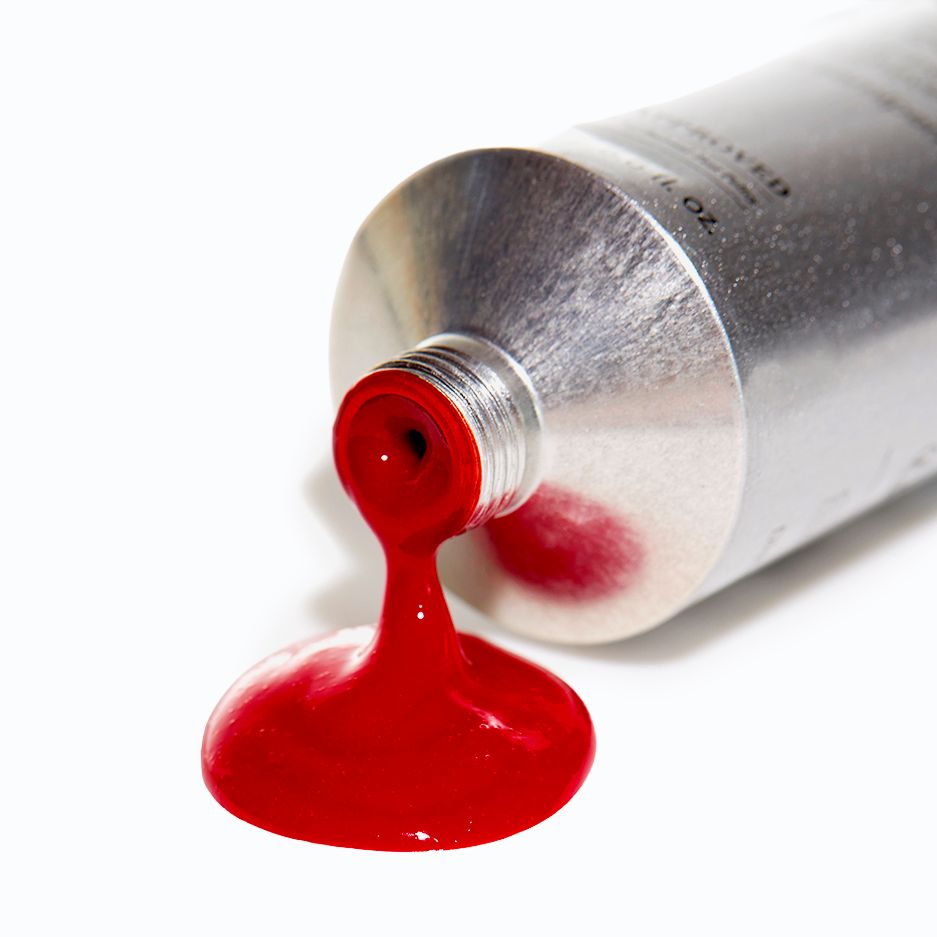 Red paint squeezed from a silver metal tube onto a white surface, creating a glossy puddle of vibrant crimson color.