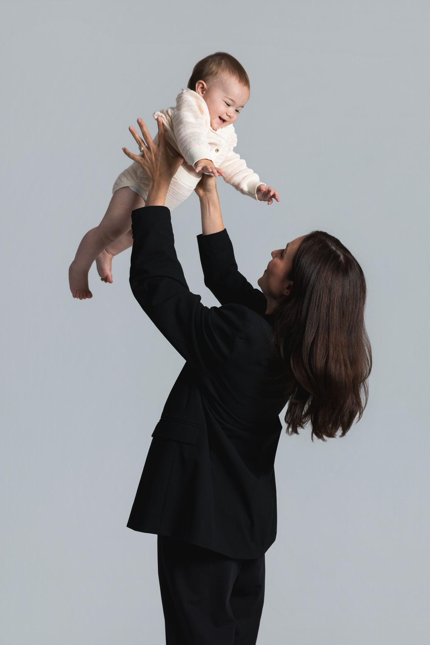 Businesswoman in black suit joyfully lifting smiling baby overhead against gray background.