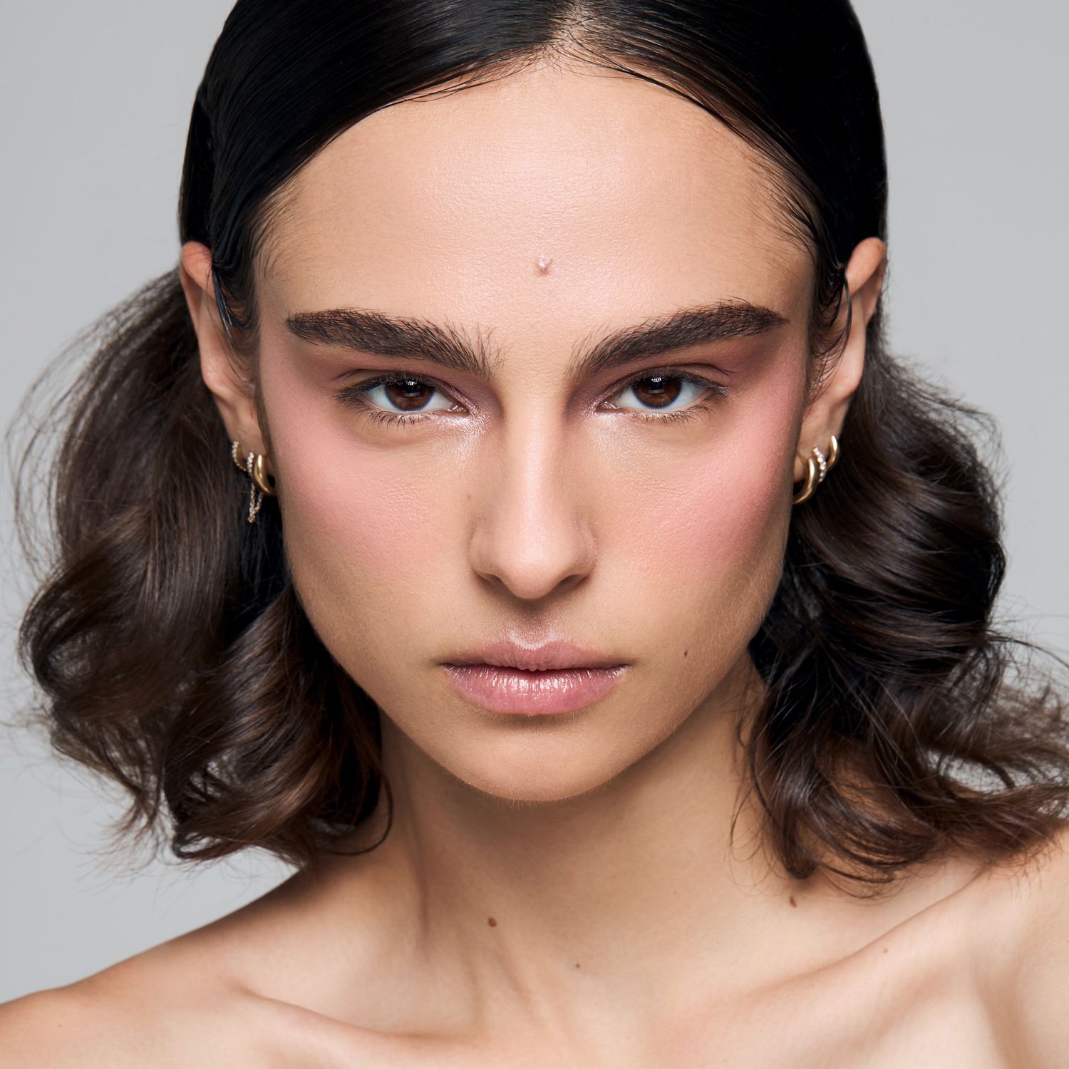 Young woman with dark wavy hair, bold brows, rosy cheeks, and gold hoop earrings in minimalist portrait.