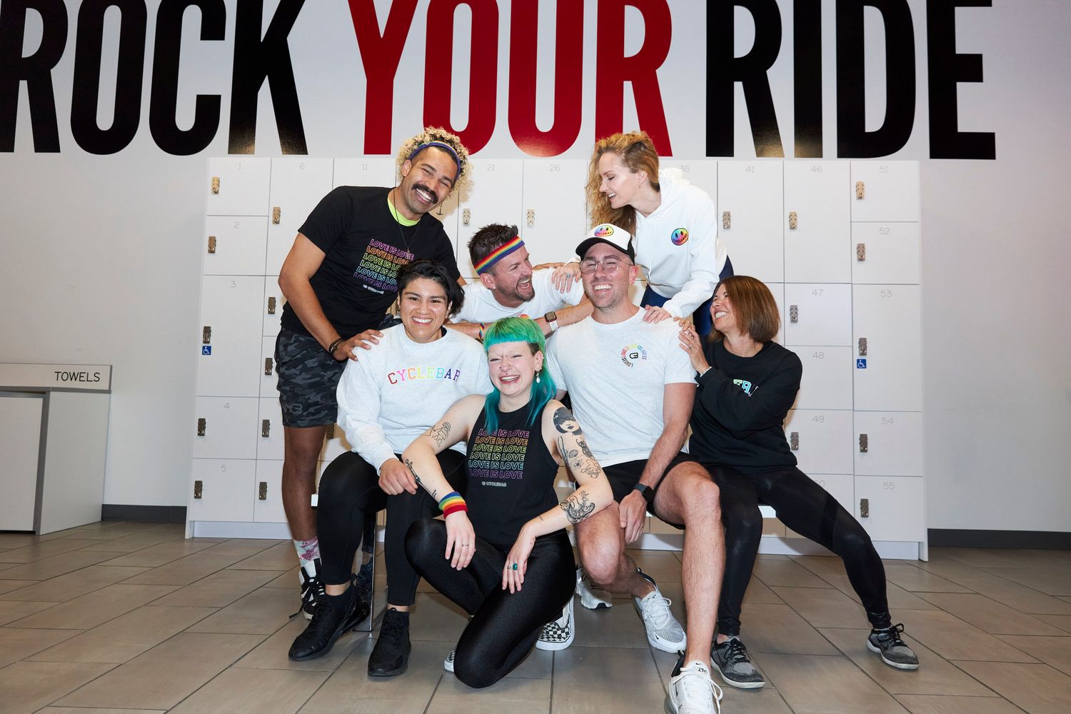 Group of friends posing and smiling in front of a Rock Your Ride wall with lockers at a cycling studio.