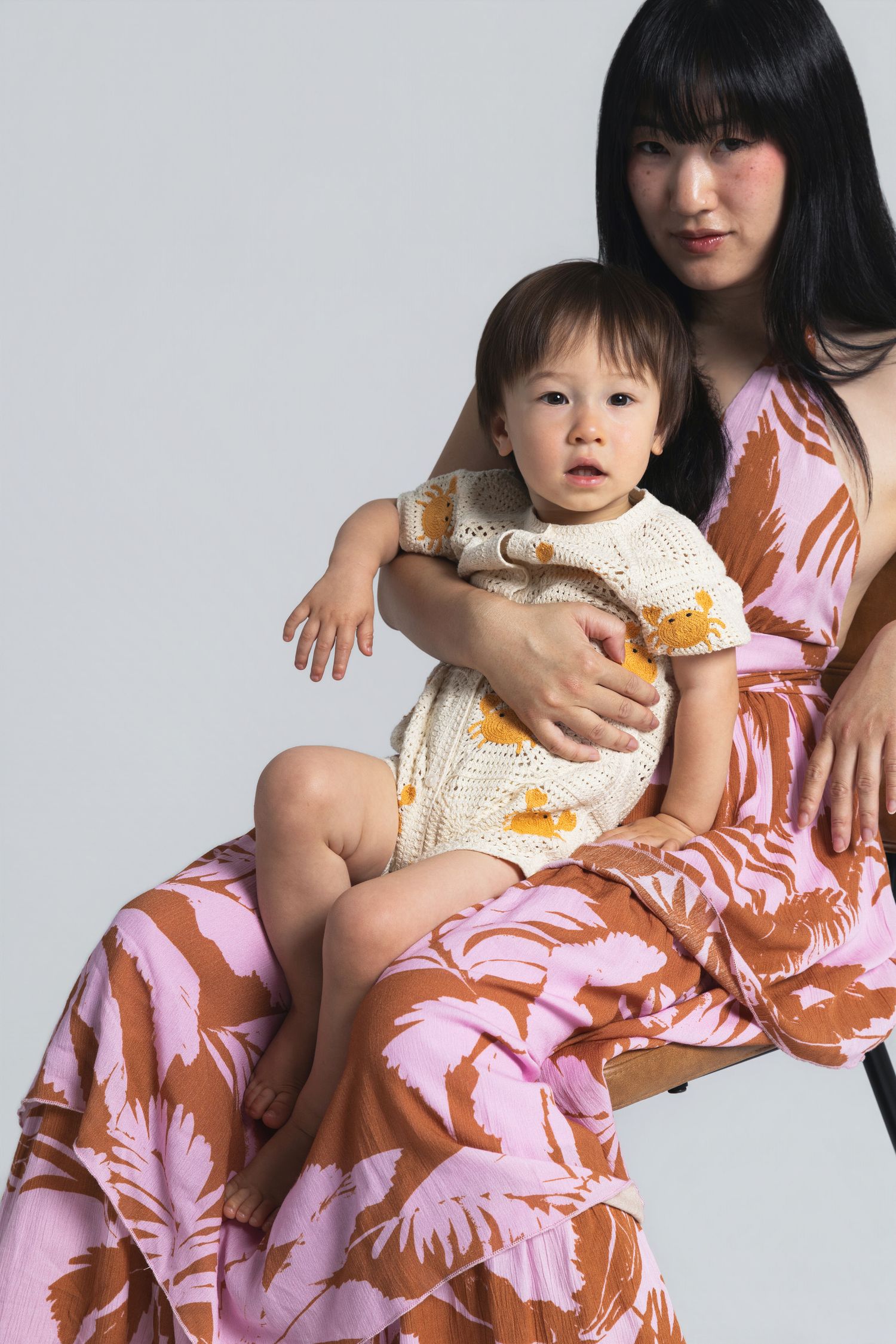 Woman in floral pink and brown dress holds baby in crochet outfit, studio portrait.