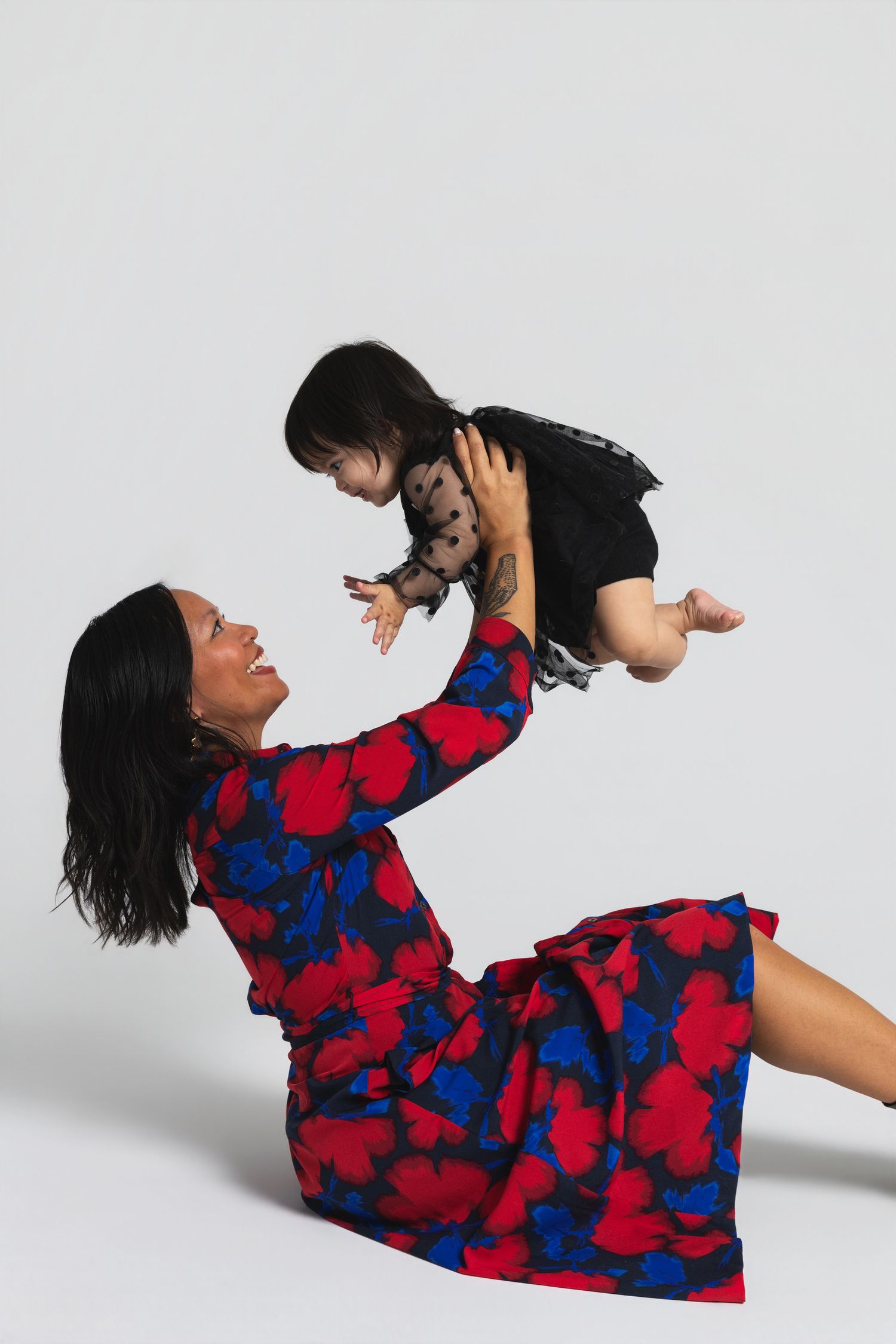 A woman in a floral dress sits on the floor, joyfully lifting a baby in a black outfit against a light background.