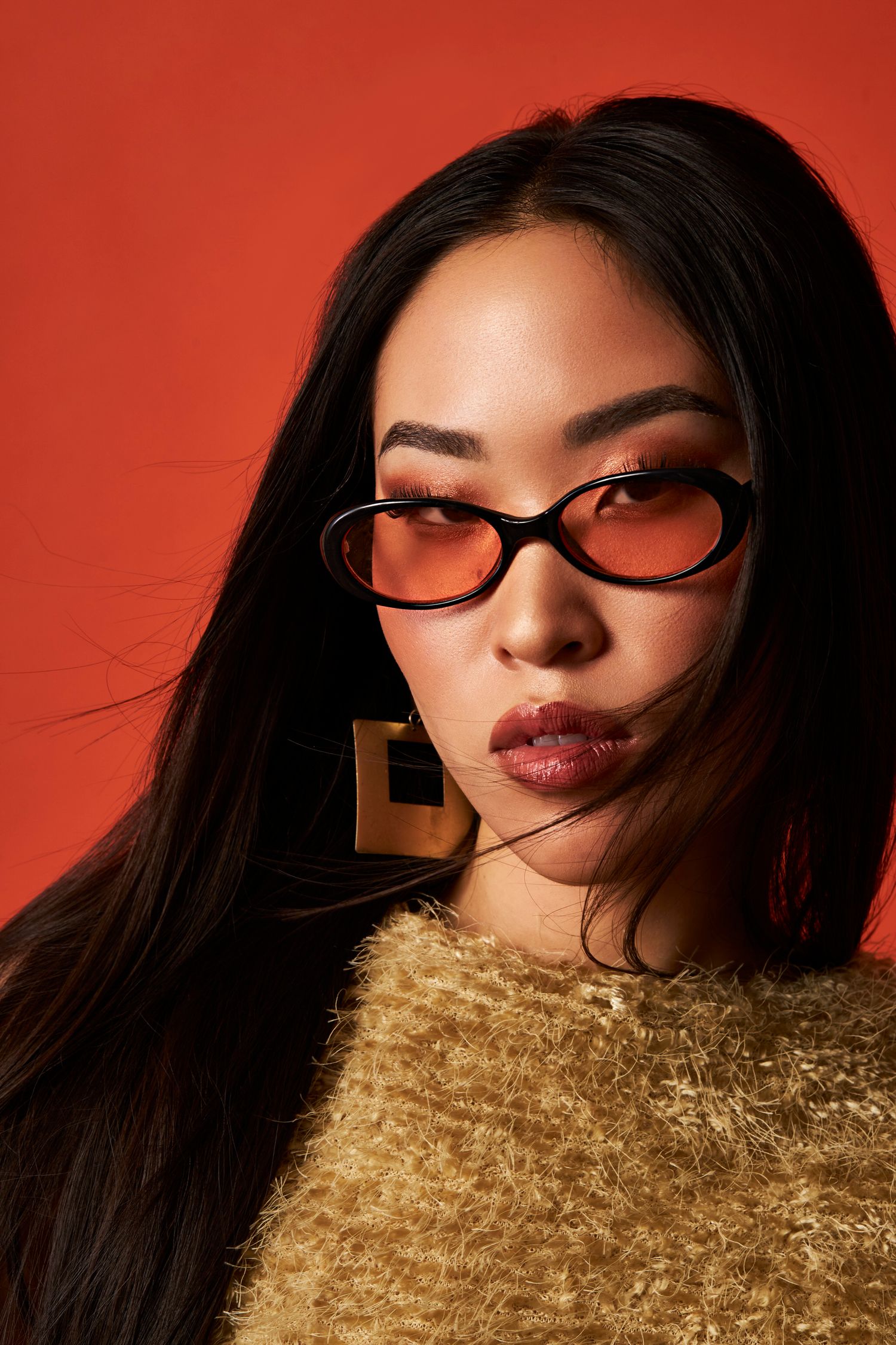 Young Asian woman in tinted oval sunglasses, gold earrings, and fuzzy sweater posing against a bold red background.