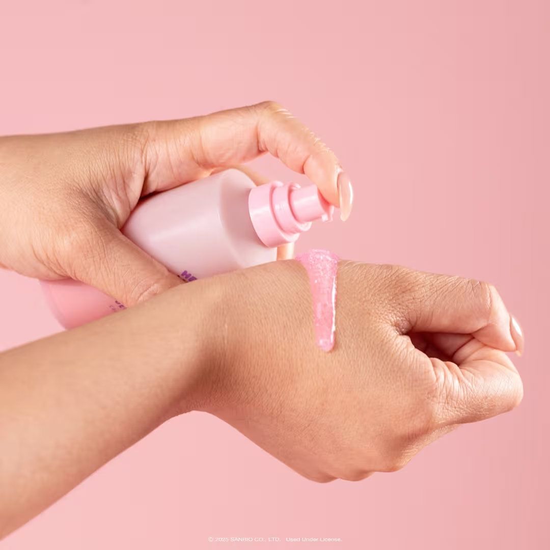 Hands dispensing pink lotion from a bottle onto wrist against a pink background.