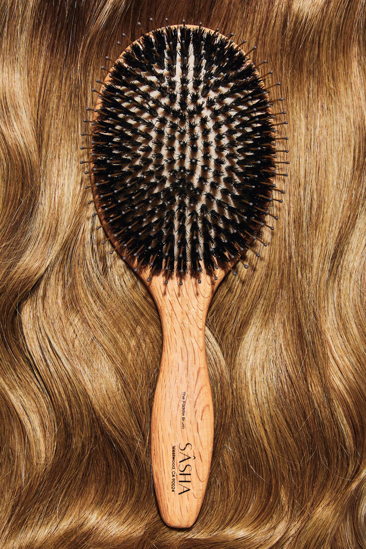 Oval wooden boar bristle paddle brush by Sasha resting on wavy golden brown hair.