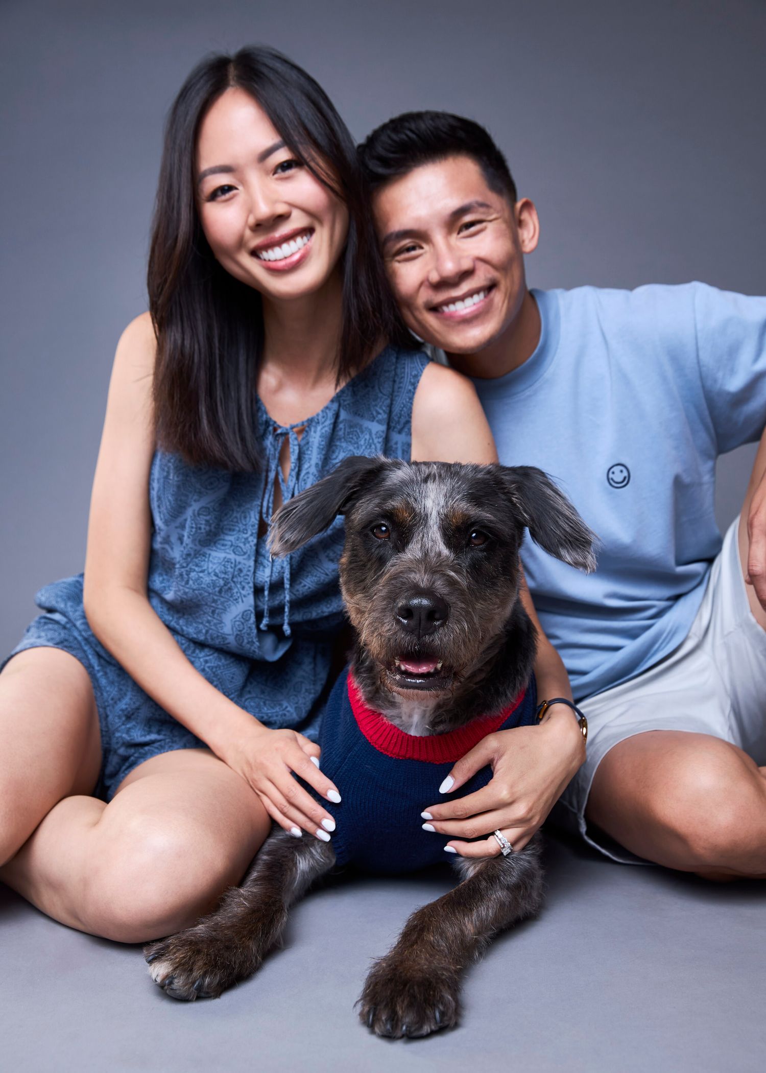 A smiling couple poses with their gray terrier mix dog wearing a red bandana in a studio portrait.
