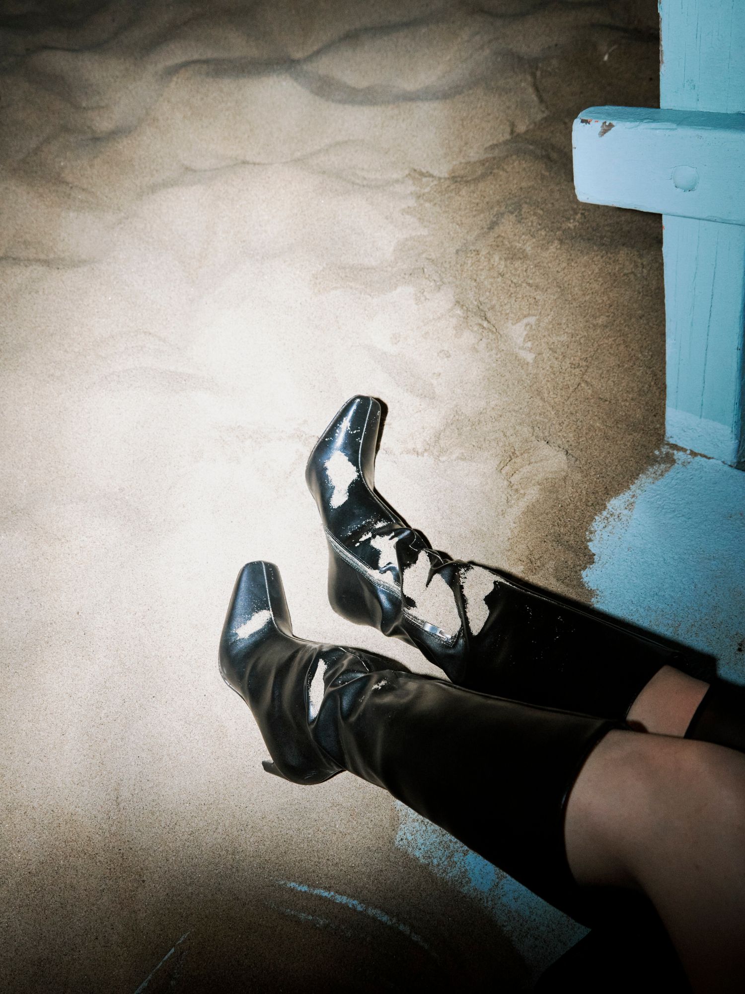 Shiny black patent leather knee-high heeled boots on sandy beach surface at night.