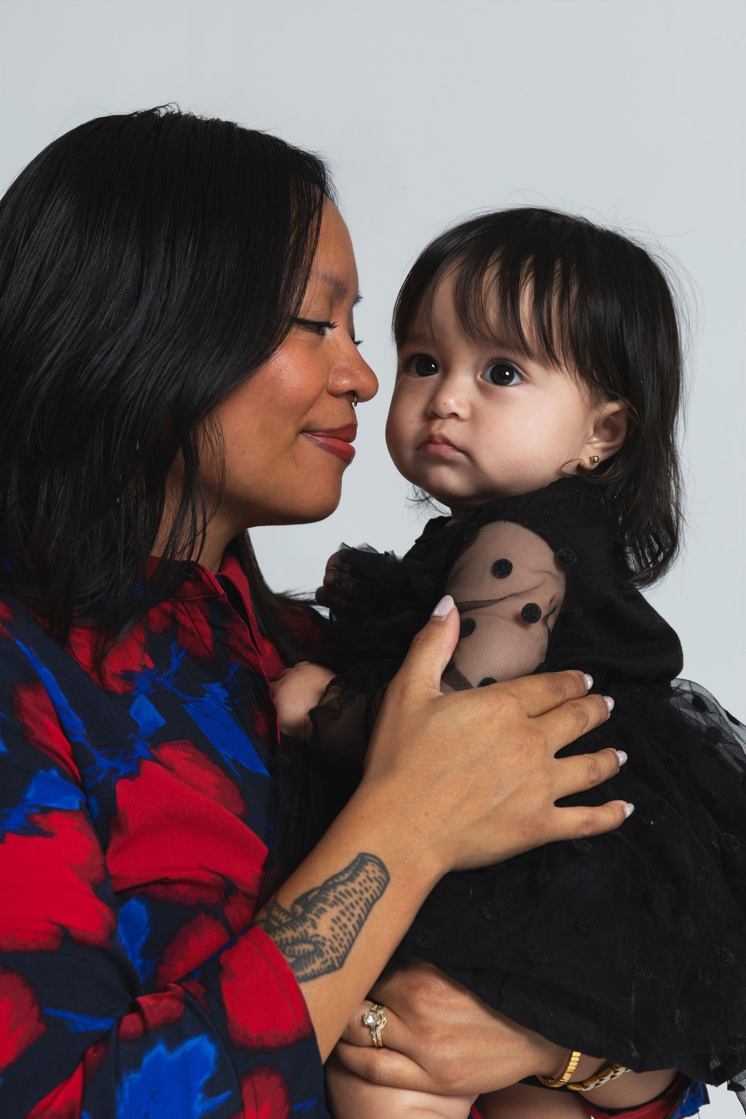 Woman in floral dress holding a baby girl in black dress, sharing a tender moment against a light background.
