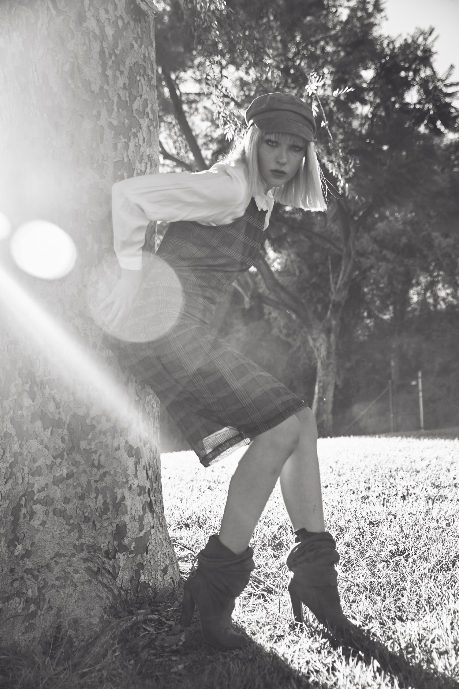 Black and white photo of a blonde woman in plaid skirt, white blouse, and cap leaning against a tree in sunlight.