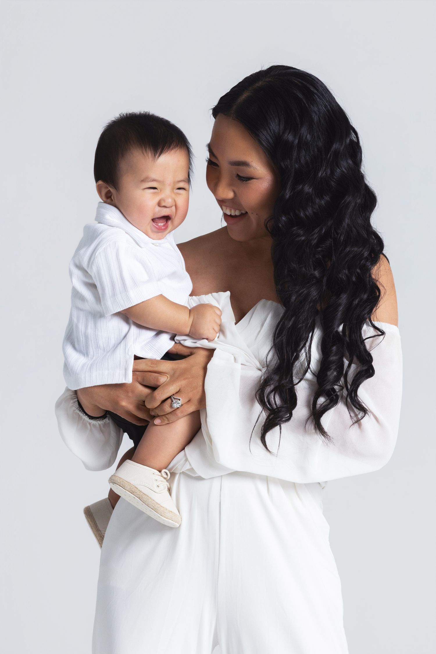 A smiling woman in white holds a laughing baby dressed in white against a light gray background.