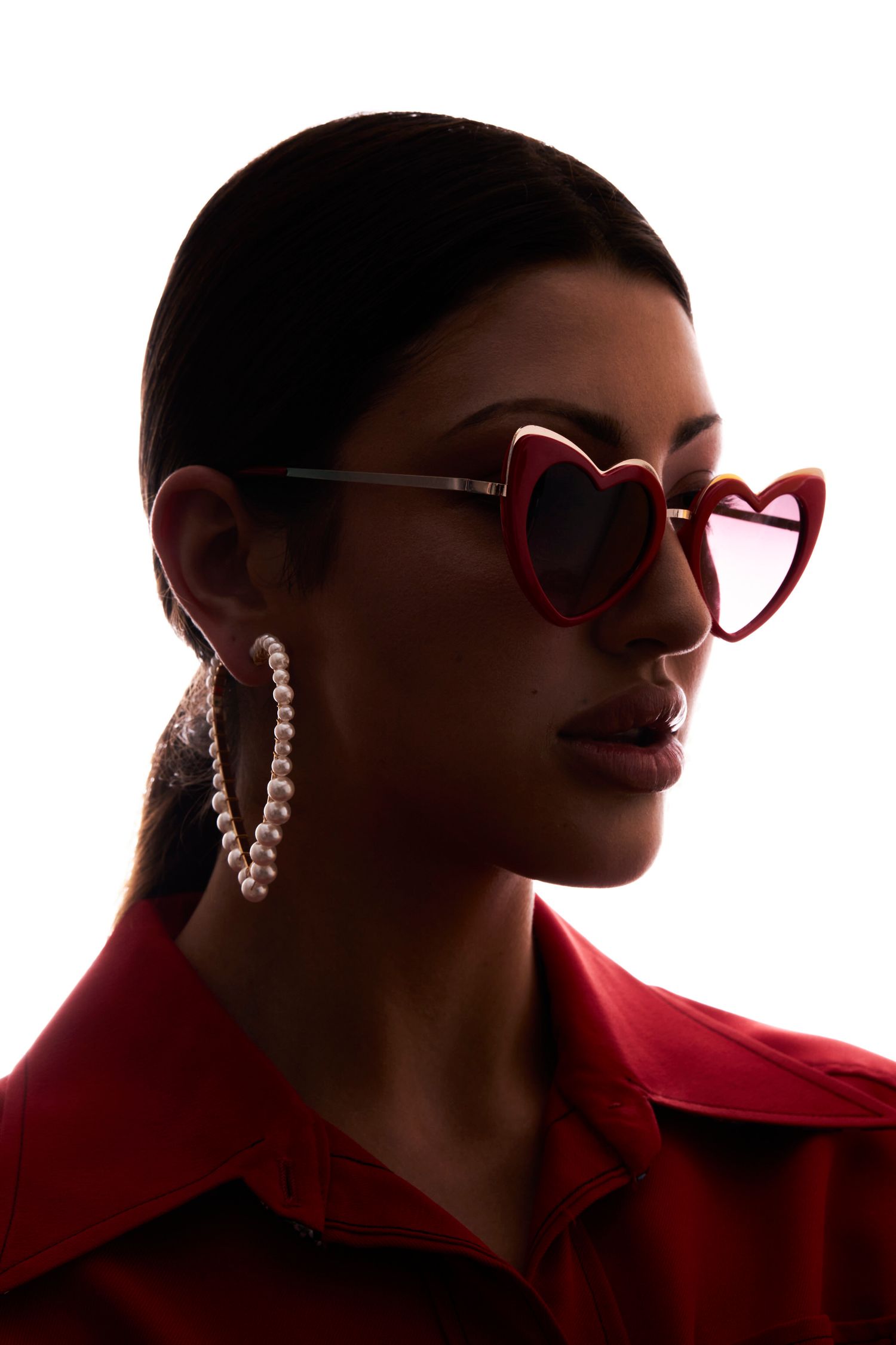 Woman in red heart-shaped sunglasses and pearl hoop earrings wearing a red jacket, posing against white background.