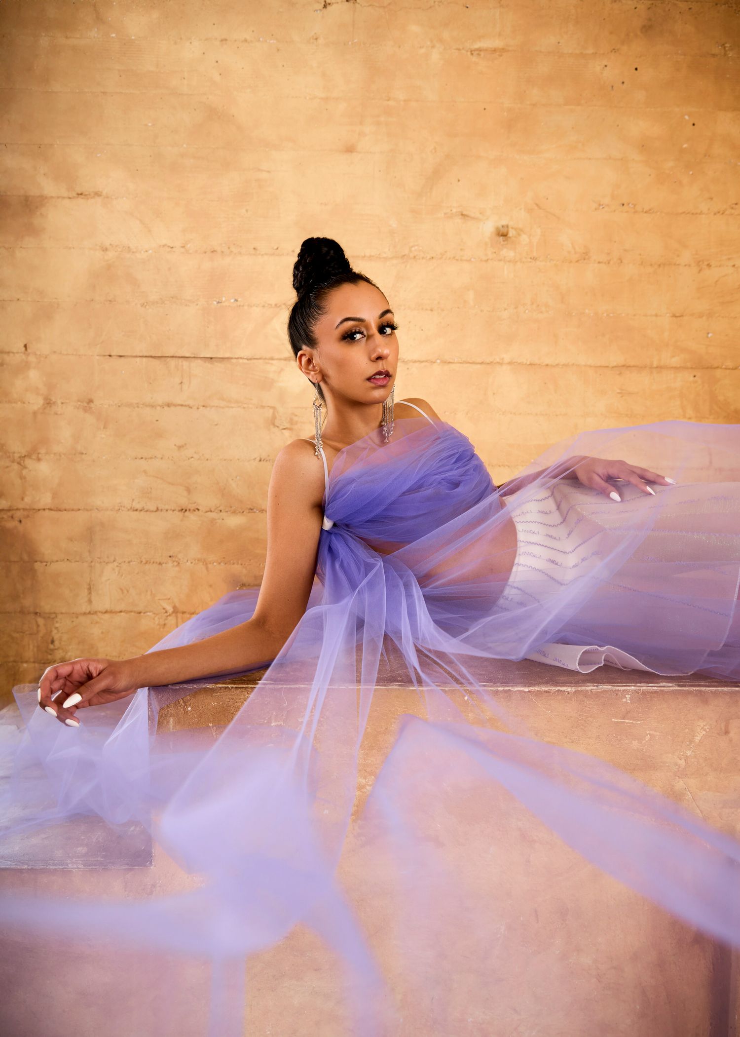 A pregnant woman in a flowing lavender tulle gown sits gracefully against a warm golden background.