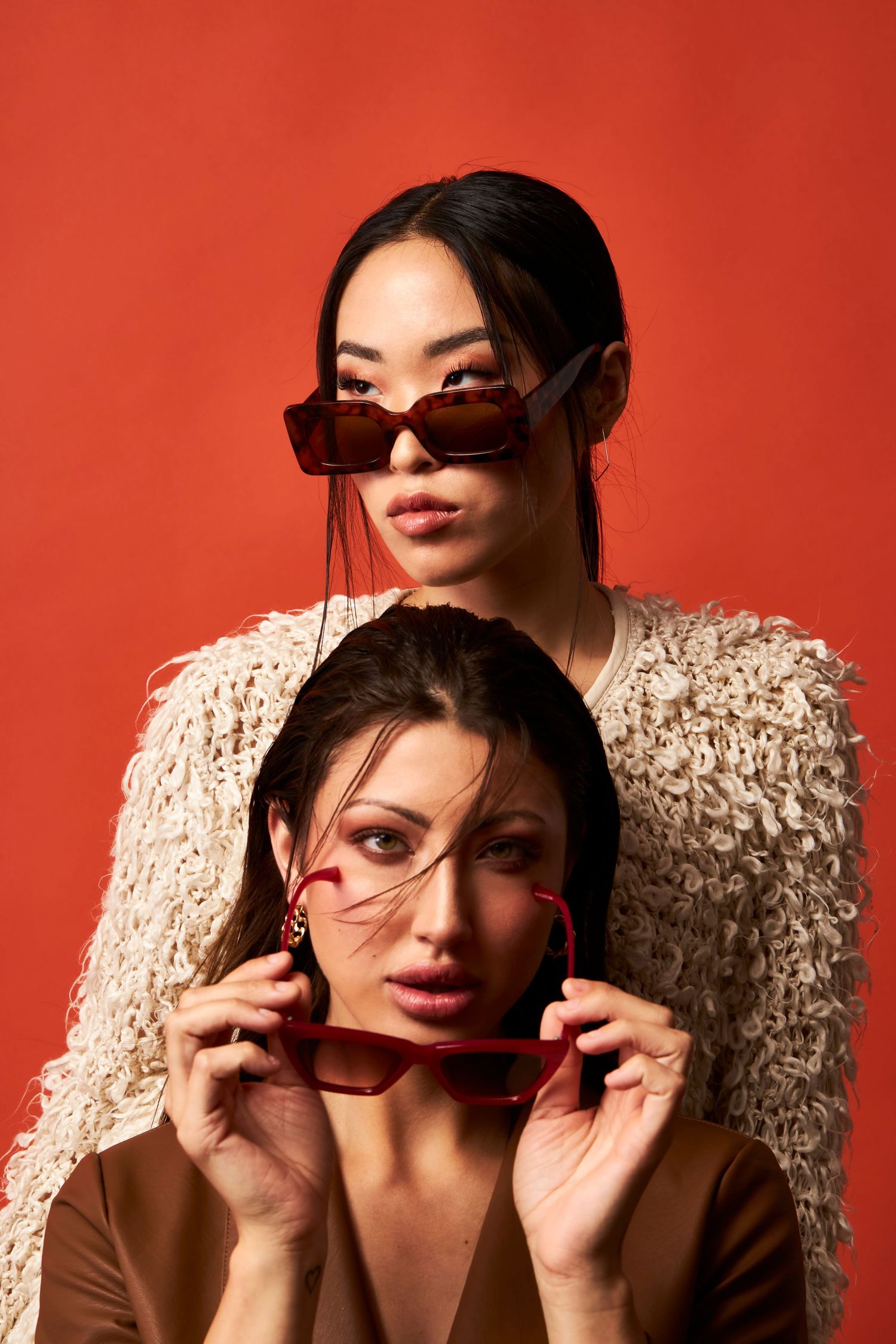 Two stylish women pose against a red background, both wearing trendy brown sunglasses and neutral-toned outfits.