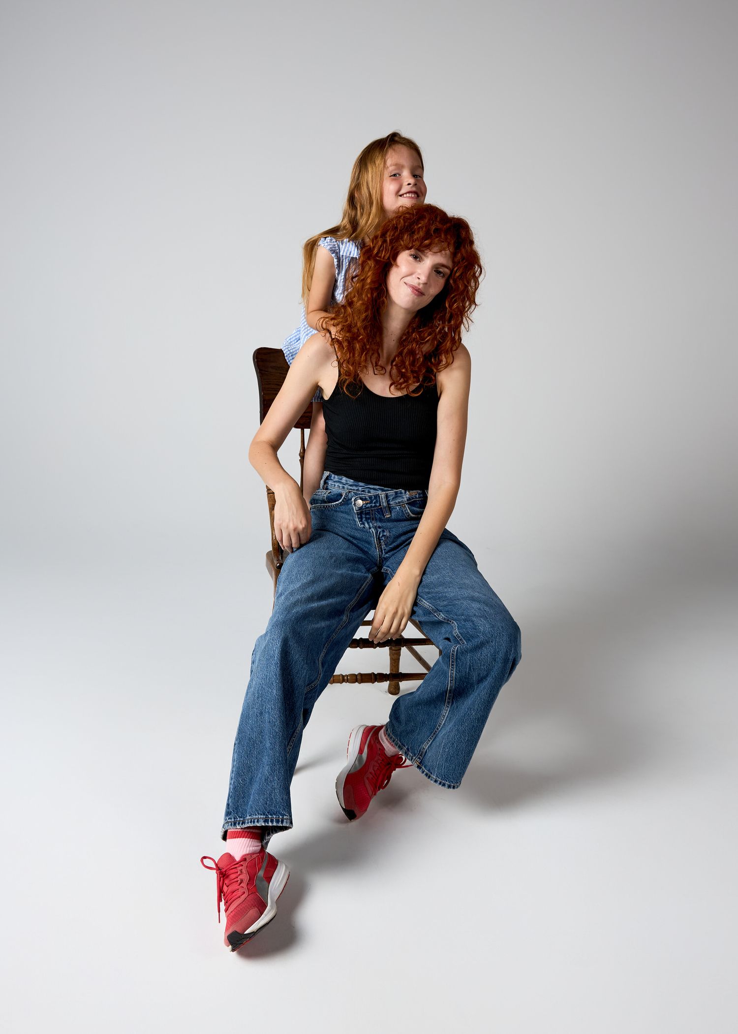 Two redhead women posing in a studio, one seated in jeans and sneakers, the other standing behind her.