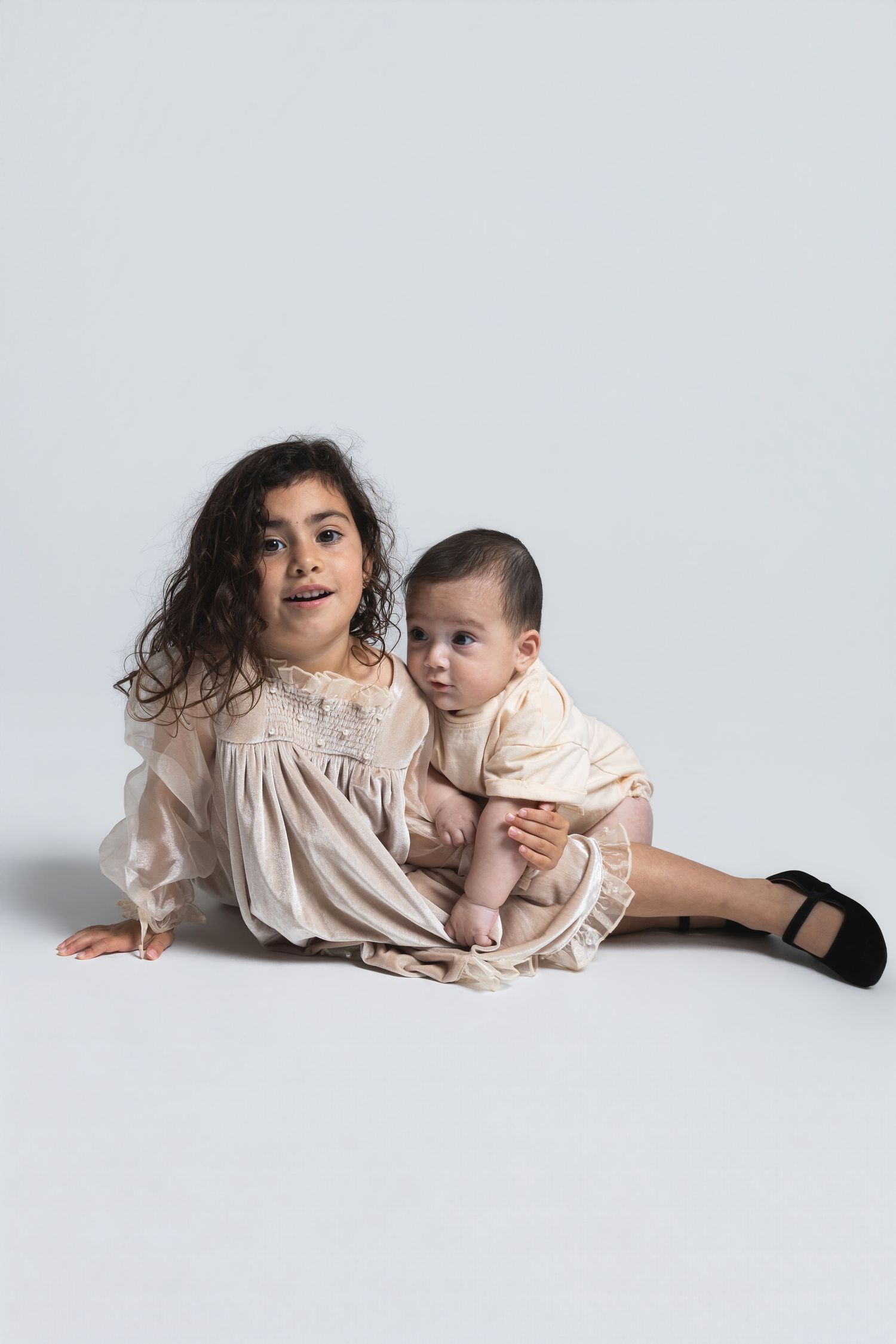 Young girl in cream dress holds baby sibling in matching outfit, both sitting on white studio floor.