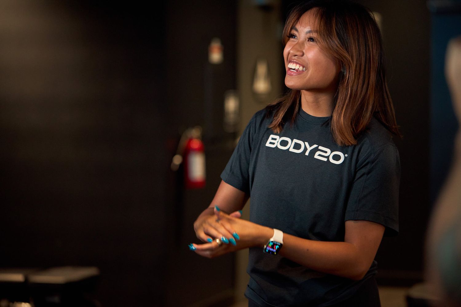 A smiling Body20 fitness trainer in a black branded t-shirt stands confidently in a gym studio.