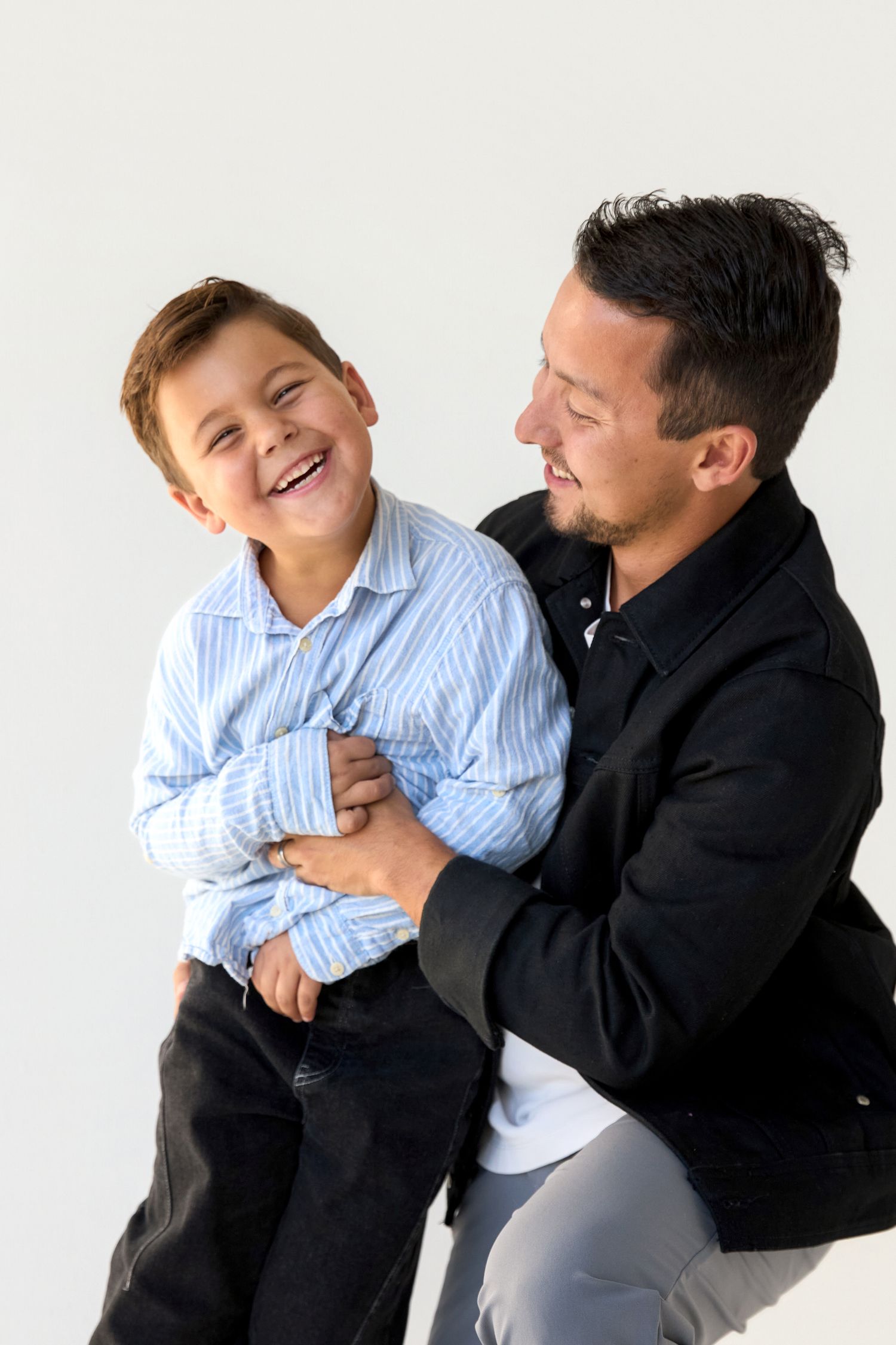 A laughing young boy in a blue striped shirt sits on a smiling man in a black jacket against a white background.