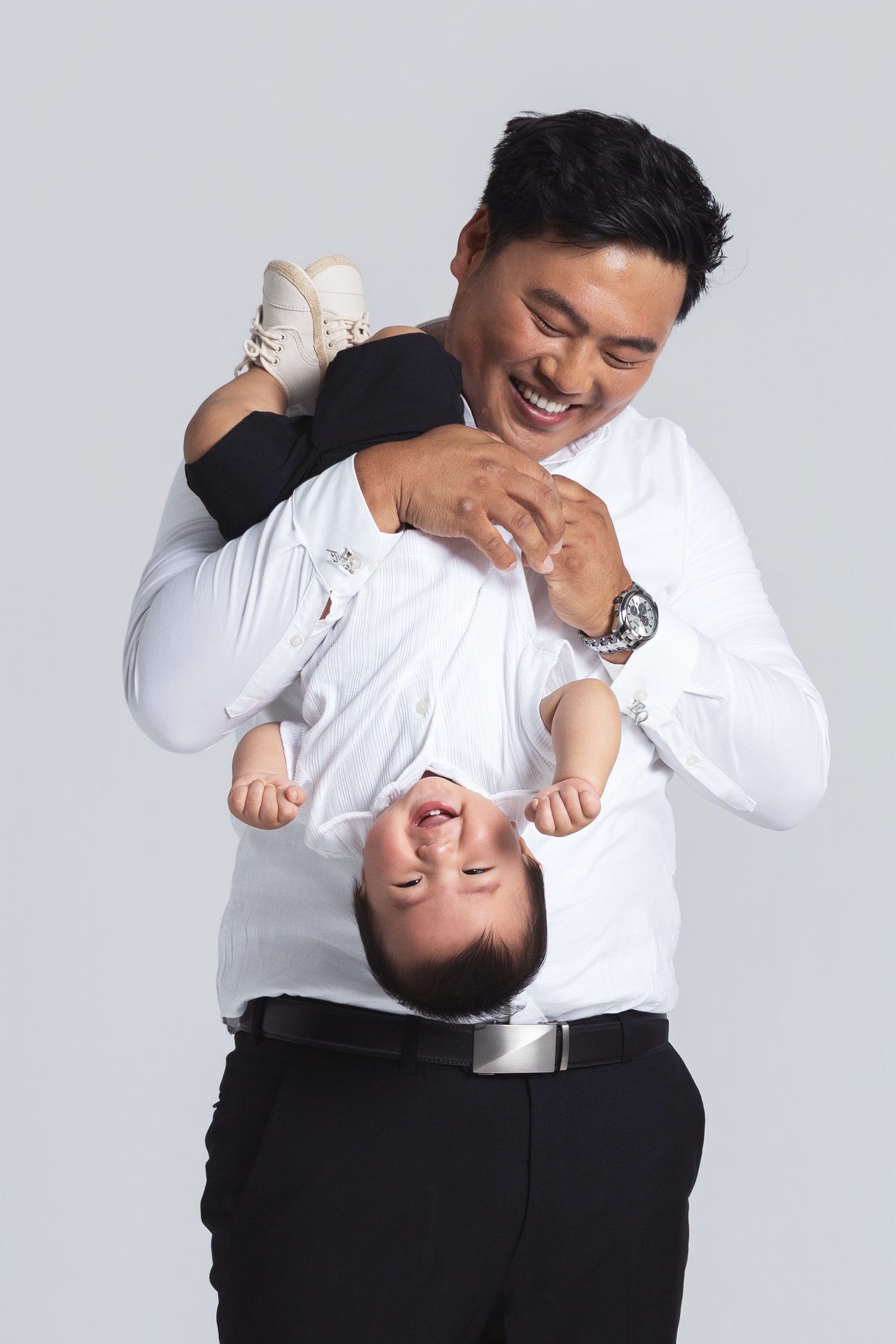 Smiling Asian father playfully holding his baby upside down against a light gray background.