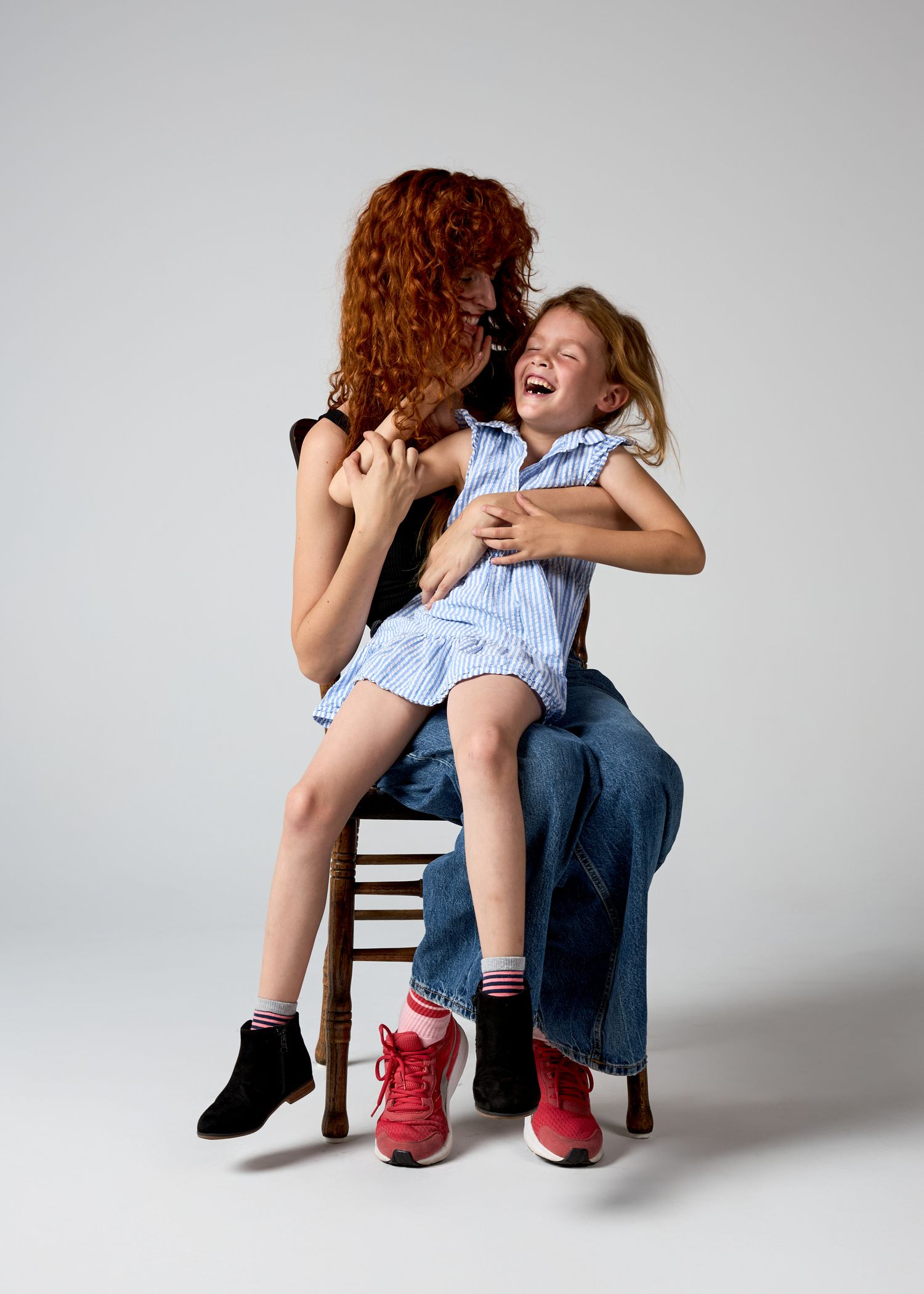 A red-haired woman tickles a laughing young girl on a chair in a bright studio photo.