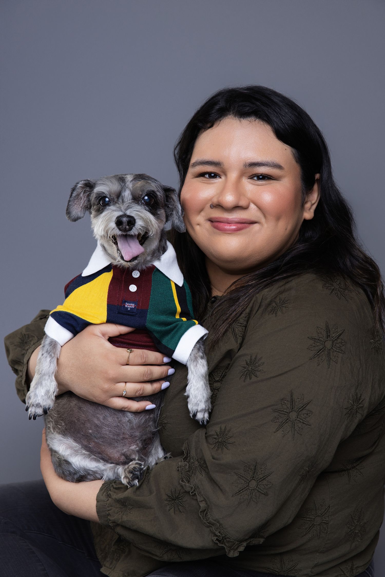 A smiling woman holds a gray Schnauzer dog wearing a colorful polo shirt, posing together against a gray background.