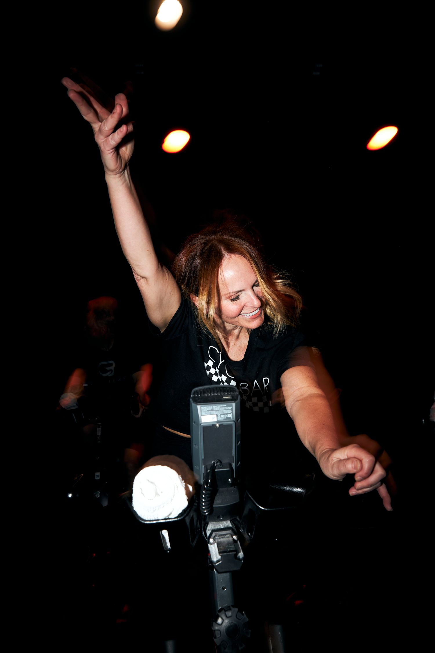 Woman raising hand and smiling on stationary bike in dark spin class studio.