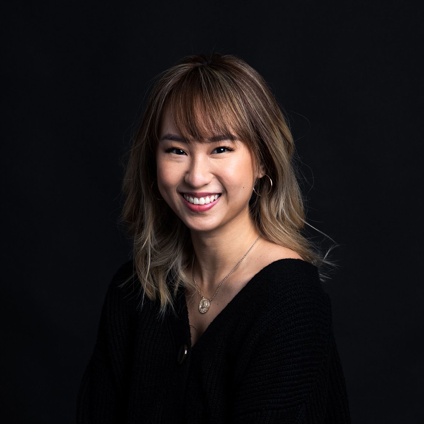 Smiling young woman with blonde highlights and bangs wearing a black top, posing against a dark studio background.