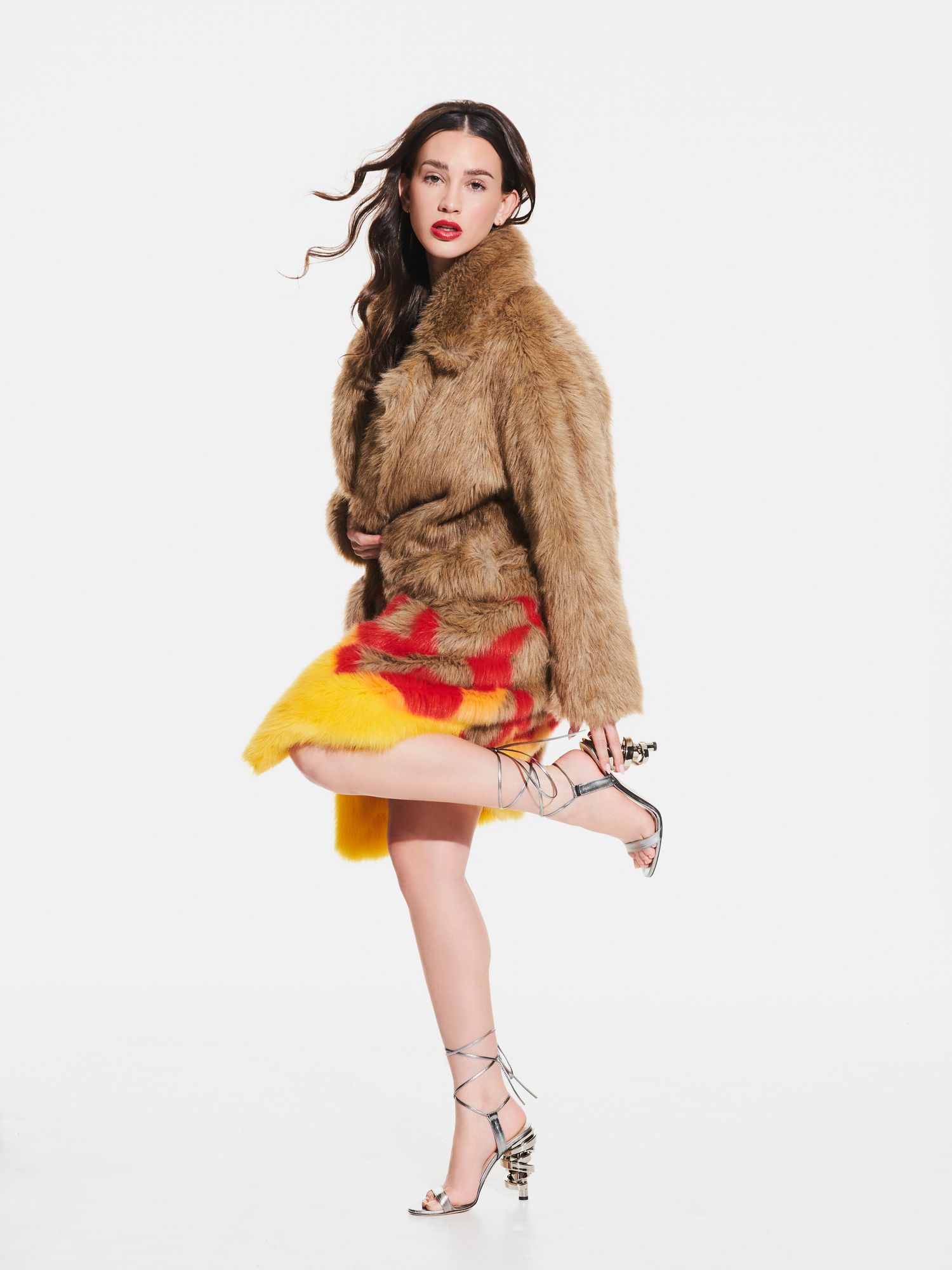 Model poses in brown faux fur coat with colorful yellow and red mini skirt, wearing strappy heels on white background.