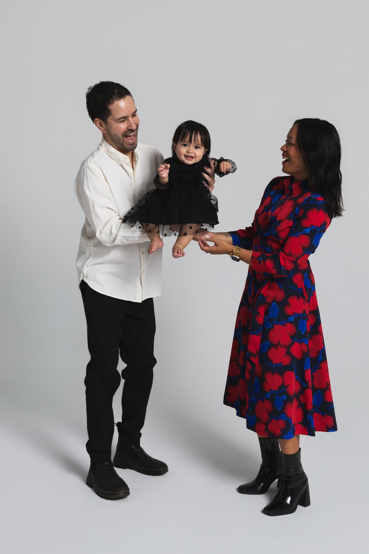 A smiling couple holds their baby girl in a black dress in a bright studio photo.