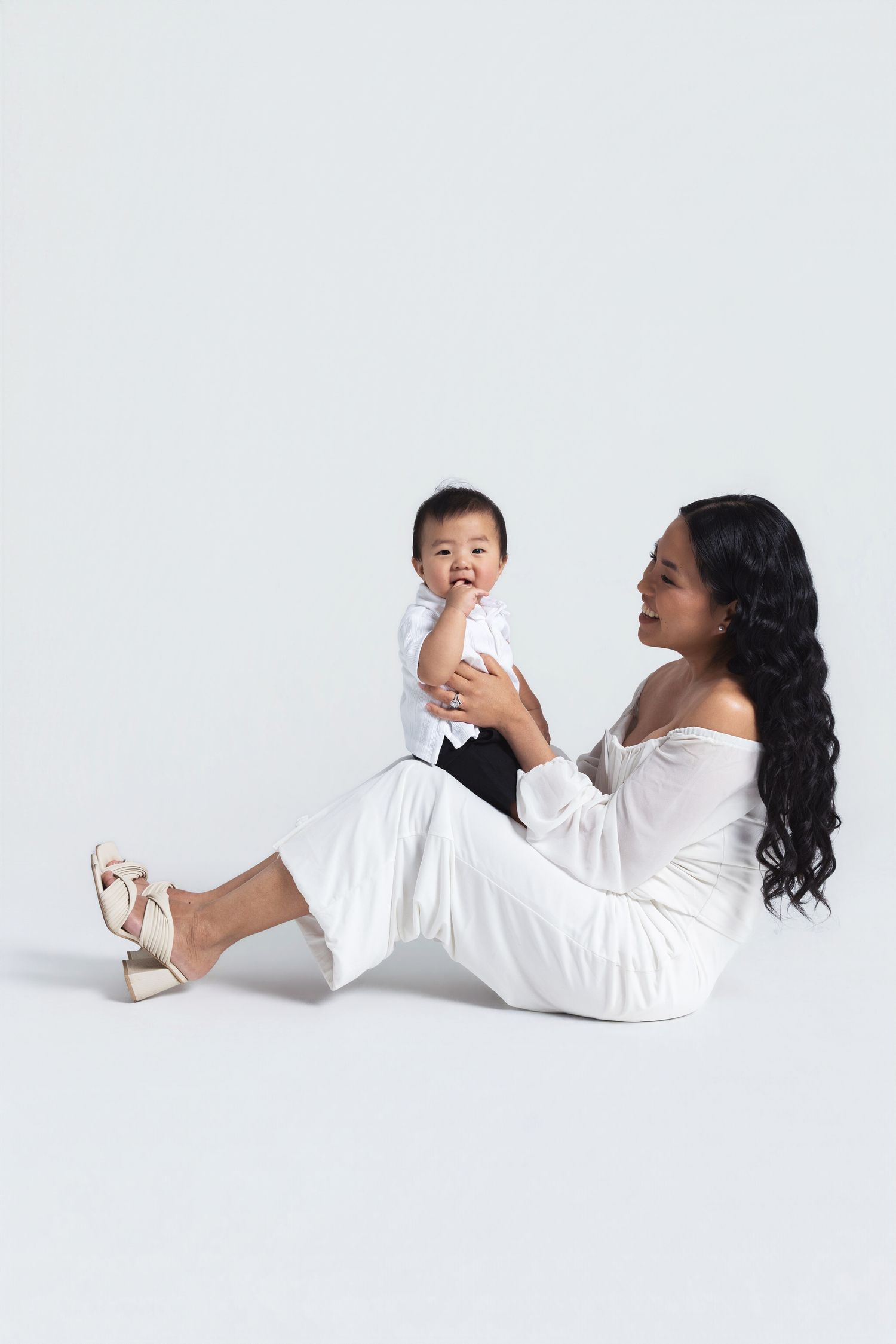 A woman in white outfit sits on floor holding her baby boy in a bright studio setting.