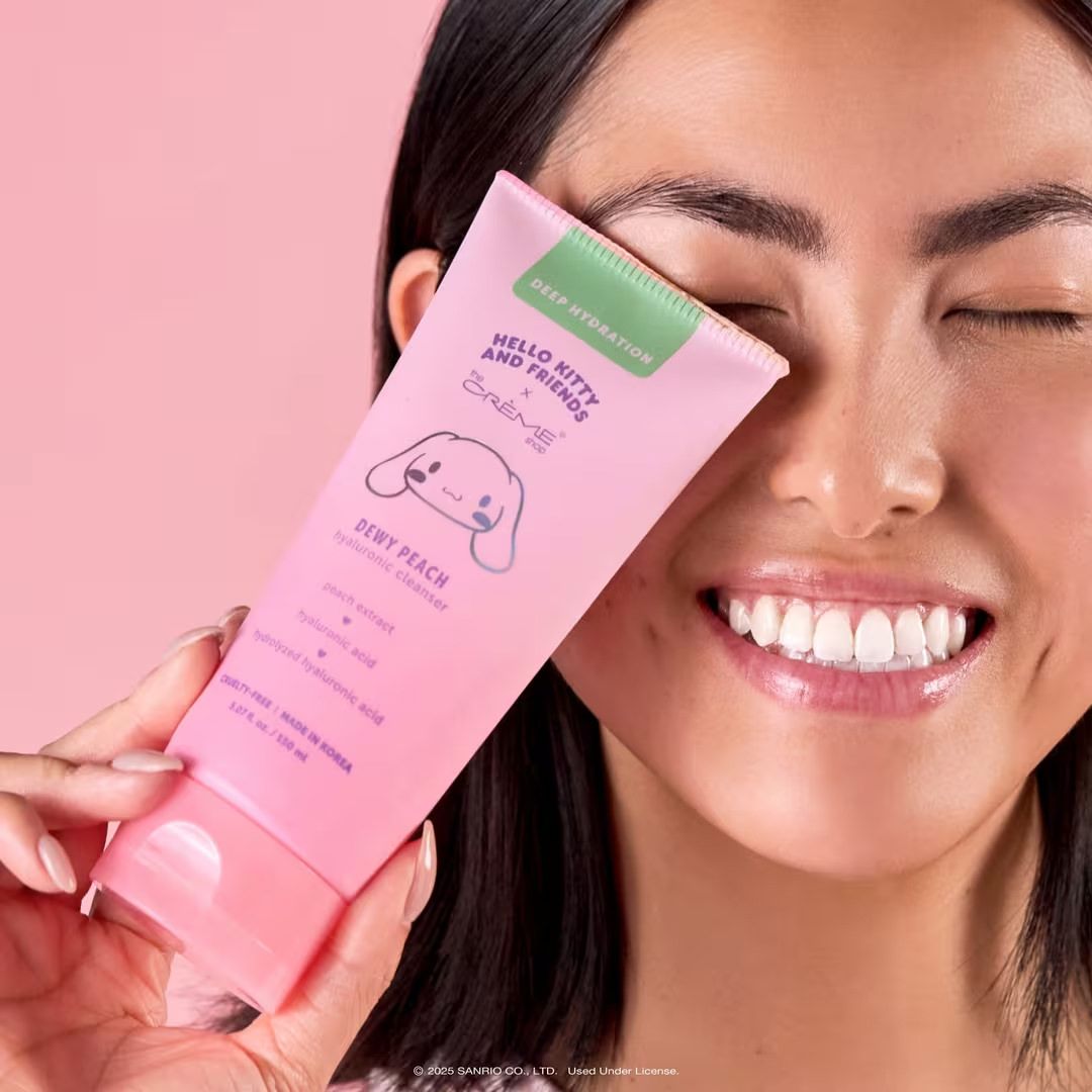 Smiling woman holding a pink Hello Kitty body primer tube in Very Peach scent against a pink background.