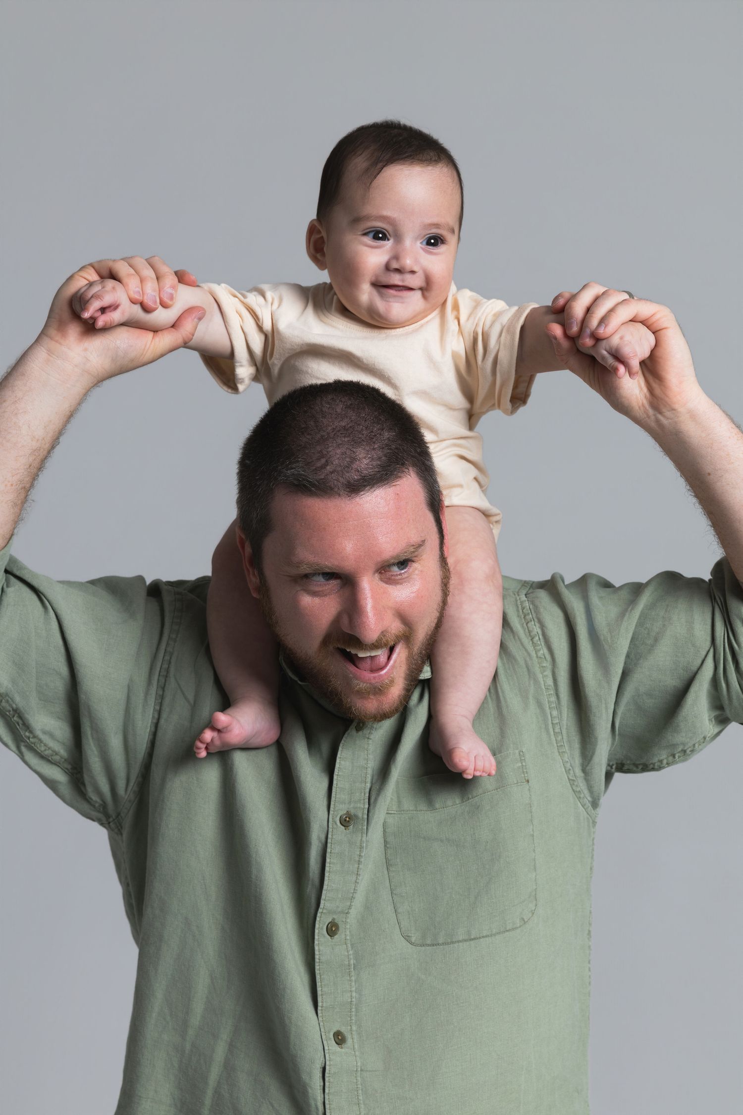 A happy baby sitting on a man's shoulders, both smiling, against a gray background.