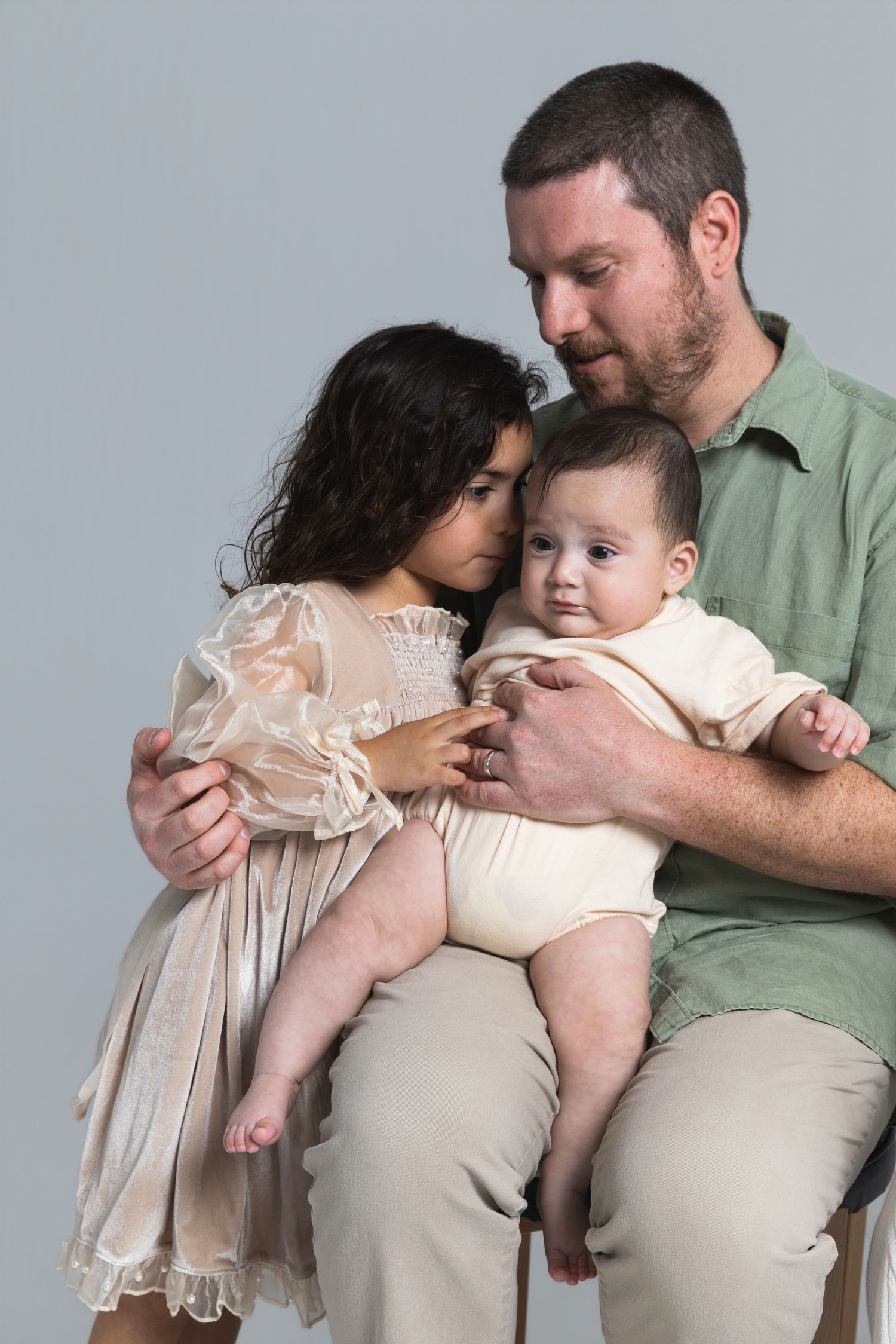 A father sits holding a baby in a onesie while a young girl in a lace dress leans in tenderly, studio portrait.