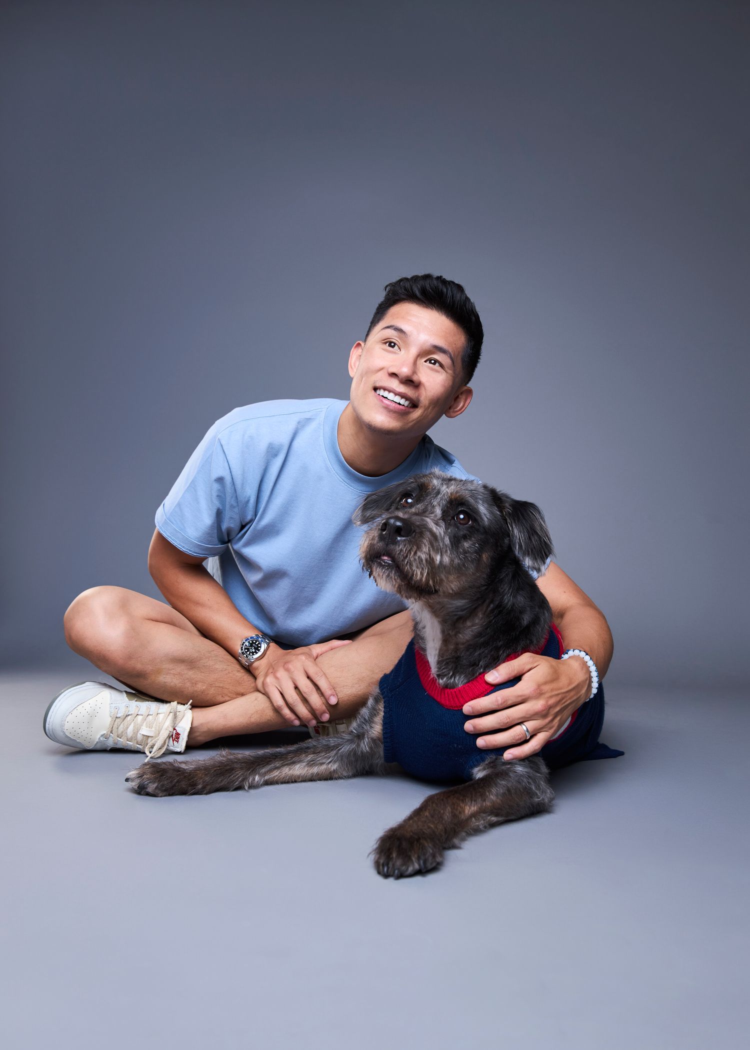 Smiling young man in blue t-shirt sitting on floor with small gray dog wearing red outfit against gray background.