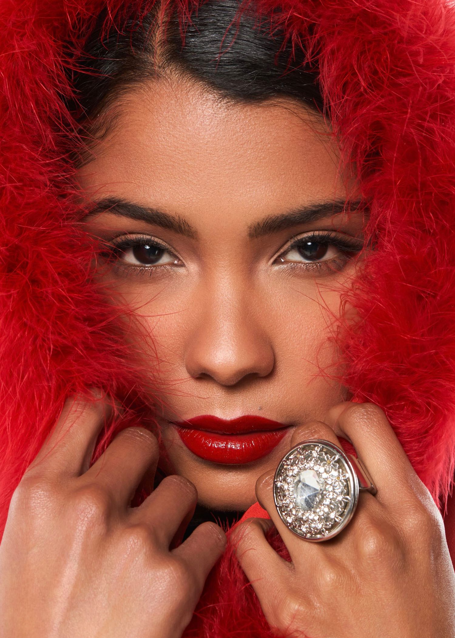 Woman in red feather boa with bold red lips and large diamond ring poses elegantly for a glamorous portrait.