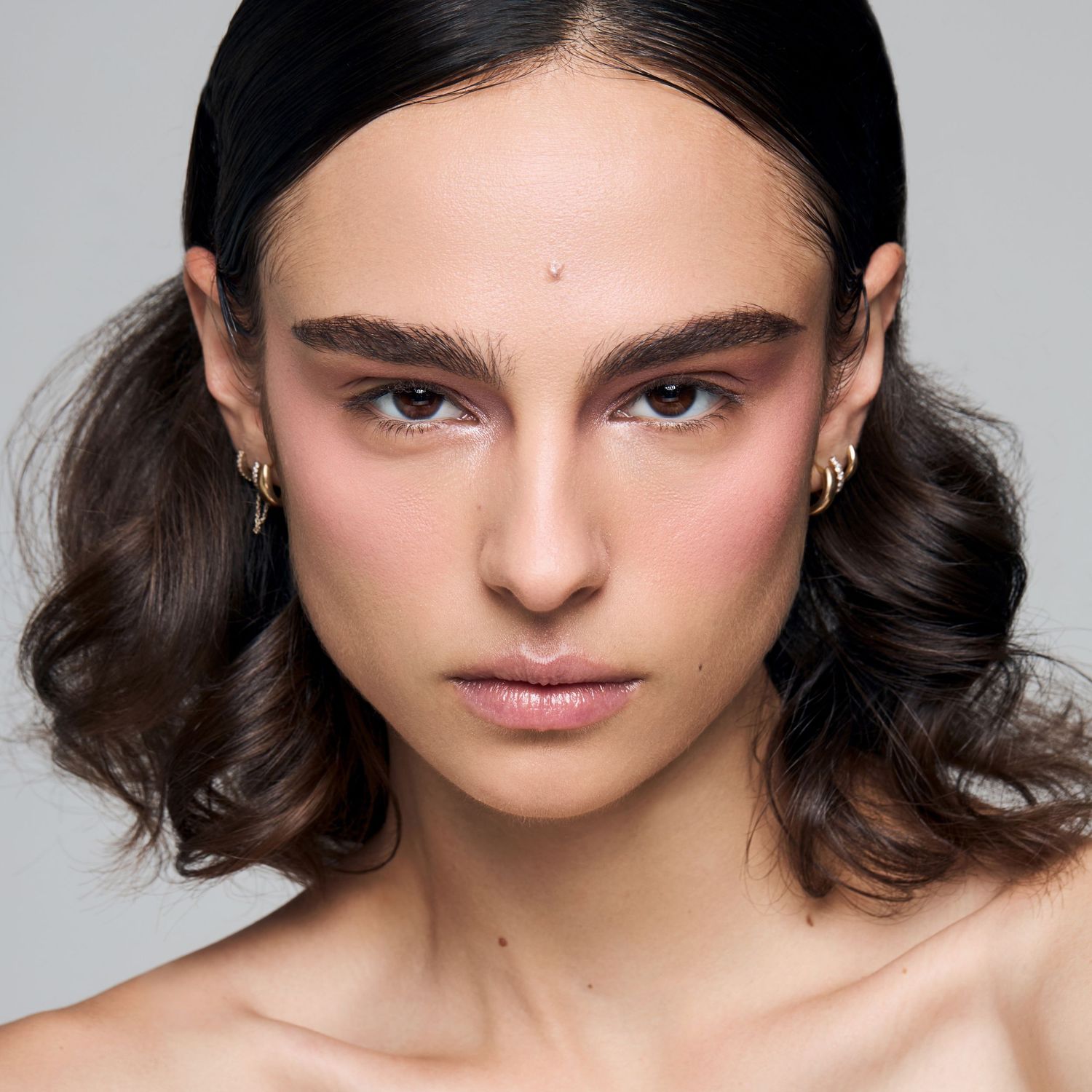 Young woman with dark wavy hair, bold brows, rosy cheeks, and gold hoop earrings in minimalist portrait.