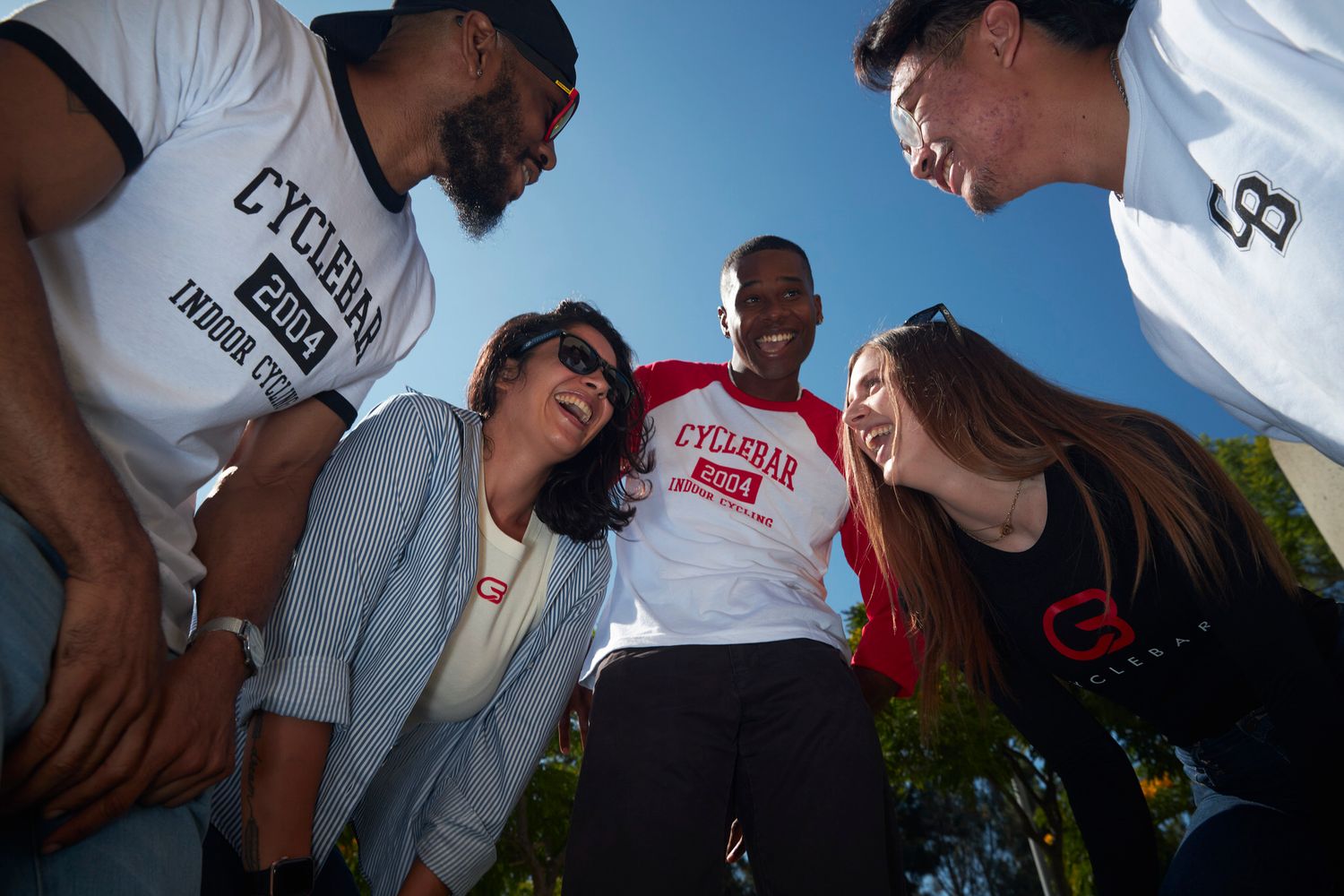 Group of smiling friends in CycleBar shirts huddle together outdoors under a blue sky.