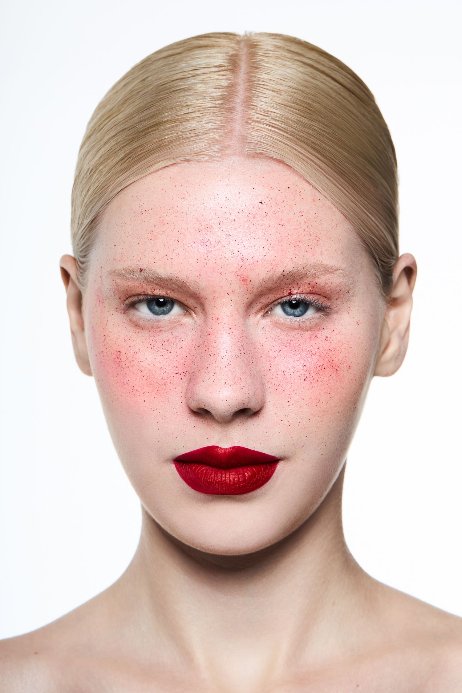 Blonde woman with blue eyes, red lips, rosy cheeks, and freckles in a minimalist beauty portrait.