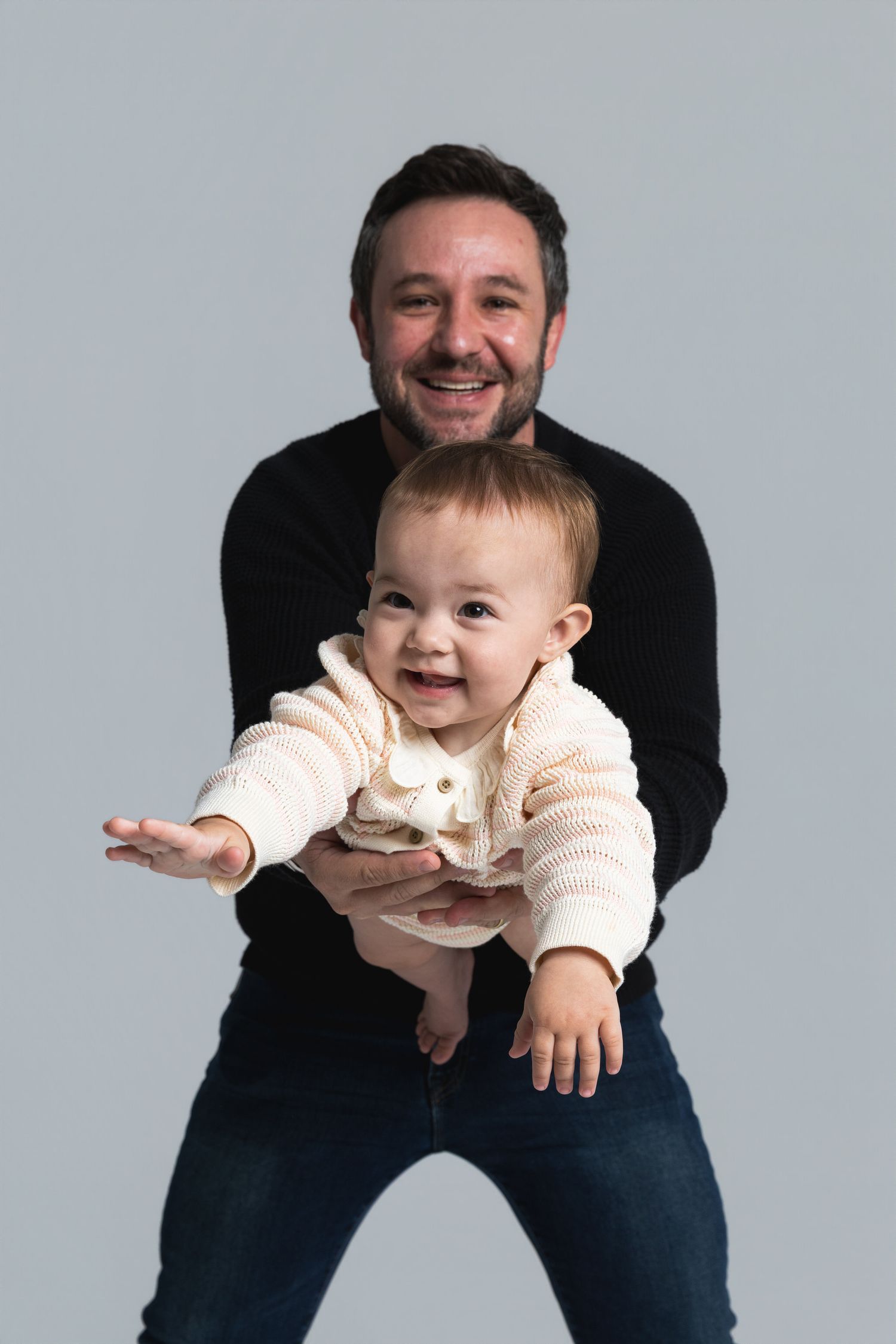 Smiling man in black sweater holding a happy baby in a cream knit outfit against a gray background.