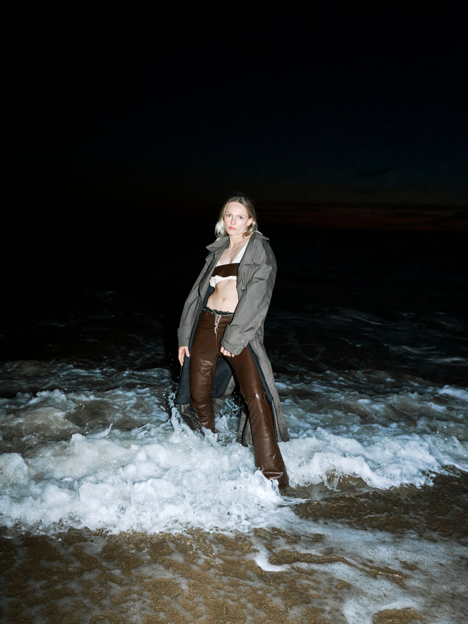 Woman in brown leather boots and coat standing in ocean waves at night on a dark beach.