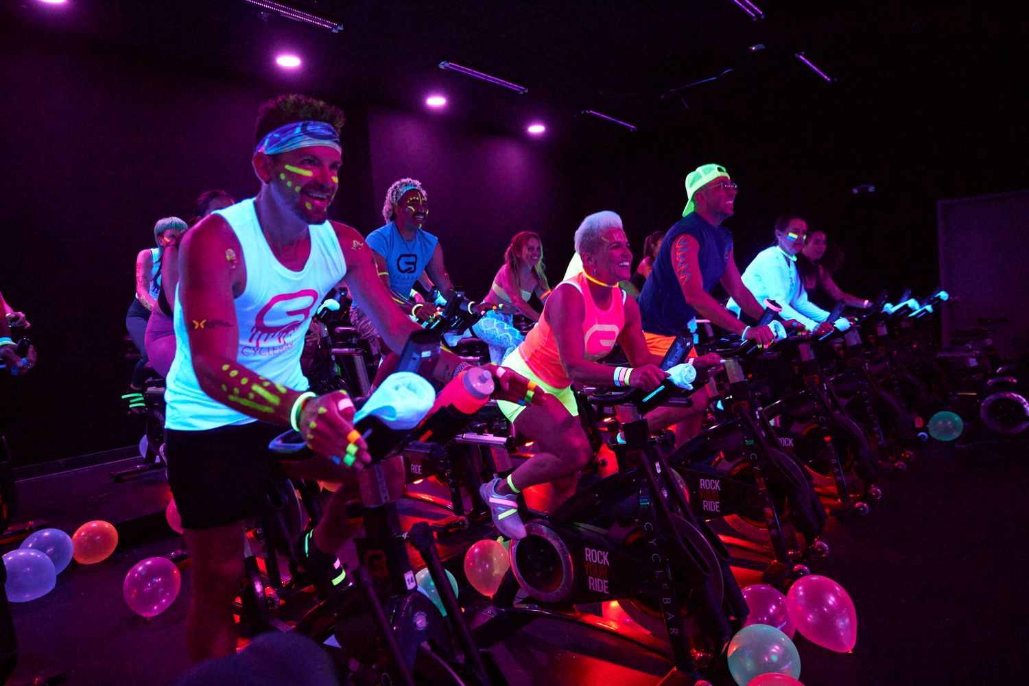 Group of cyclists in neon outfits ride stationary bikes in a dark, vibrant spin class with glowing lights.