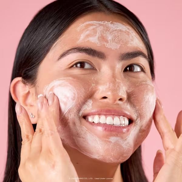 Smiling young woman applying foamy facial cleanser to her face against a pink background.