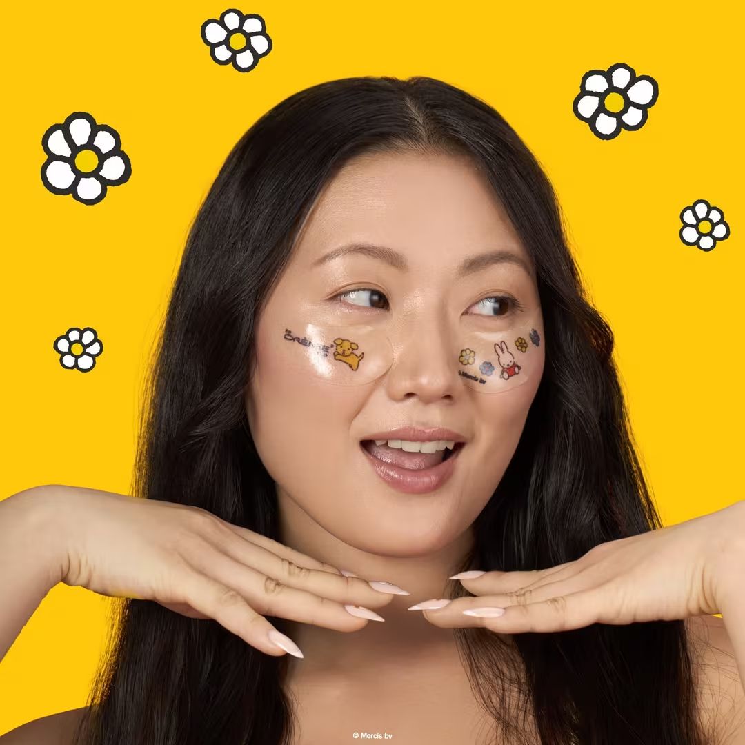 A young woman with face stickers poses against a bright yellow background with daisy illustrations, hands framing her face.