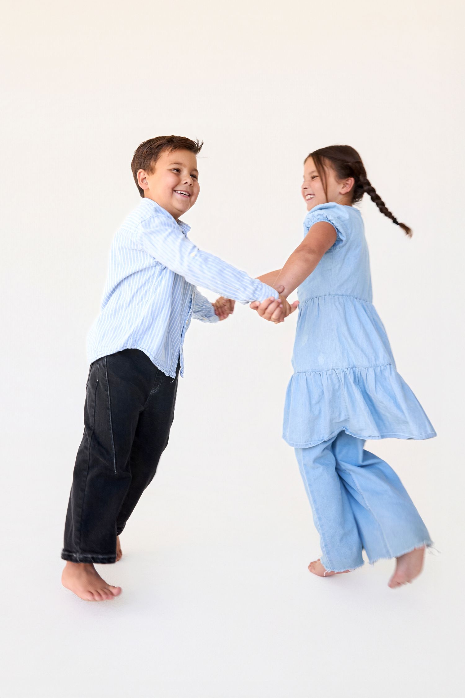 Two barefoot children in blue outfits hold hands and spin together, smiling joyfully against a white background.
