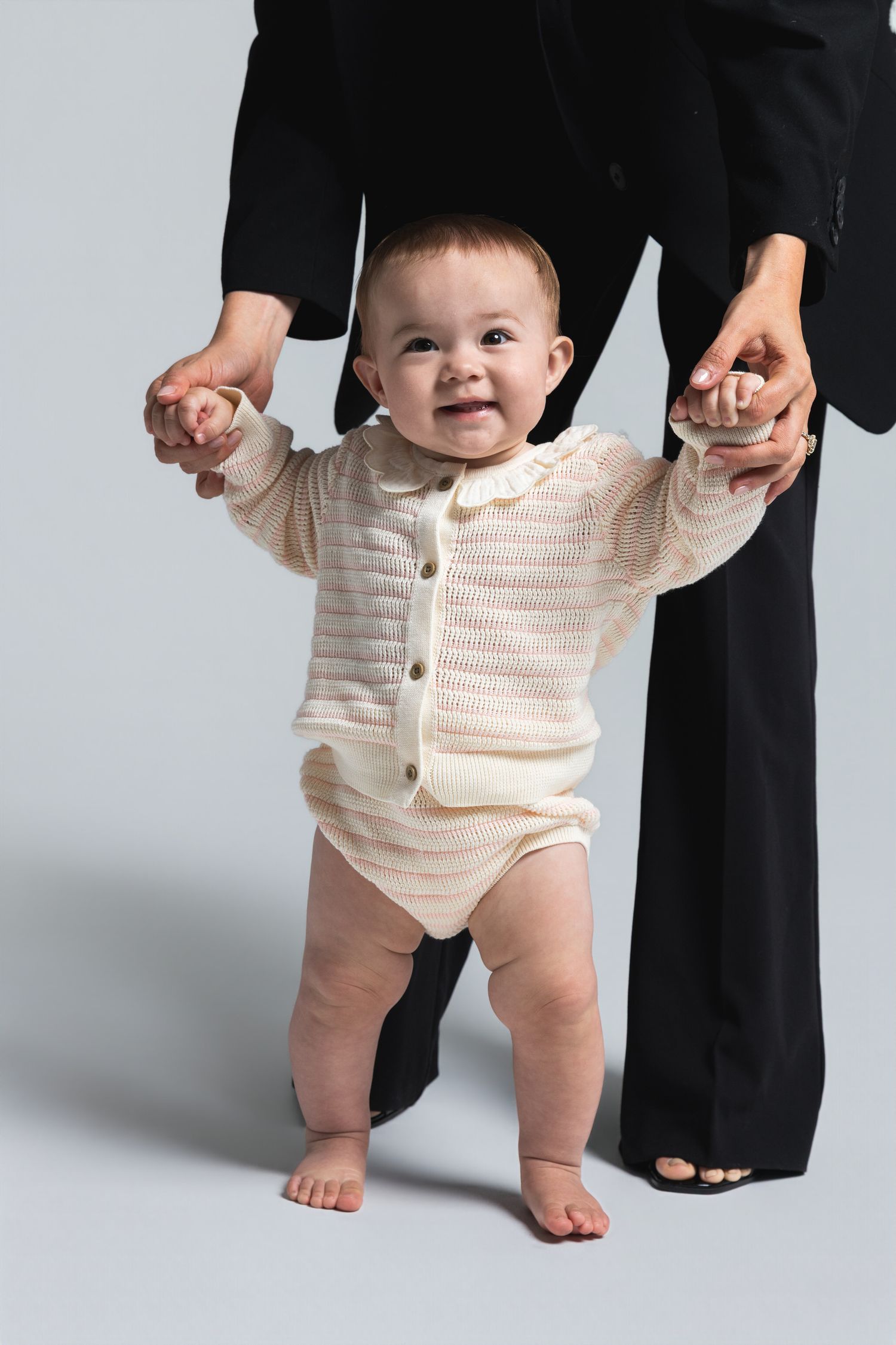 Smiling baby in cream knit cardigan learning to walk with adult support on gray background.
